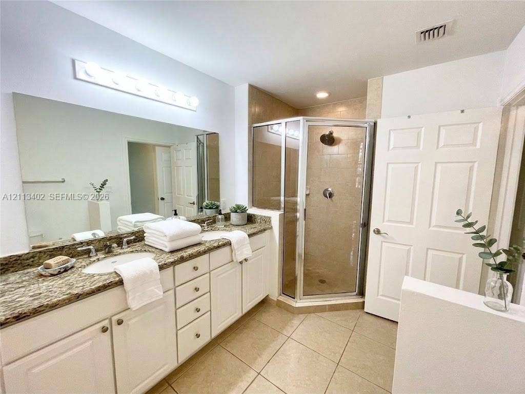3001 NE 185th Street, #207, Aventura, FL 33180 Bathroom featuring a dual-sink vanity with stone-surface countertops, expansive wall-mounted mirror, corner glass-enclosed shower, and tile flooring