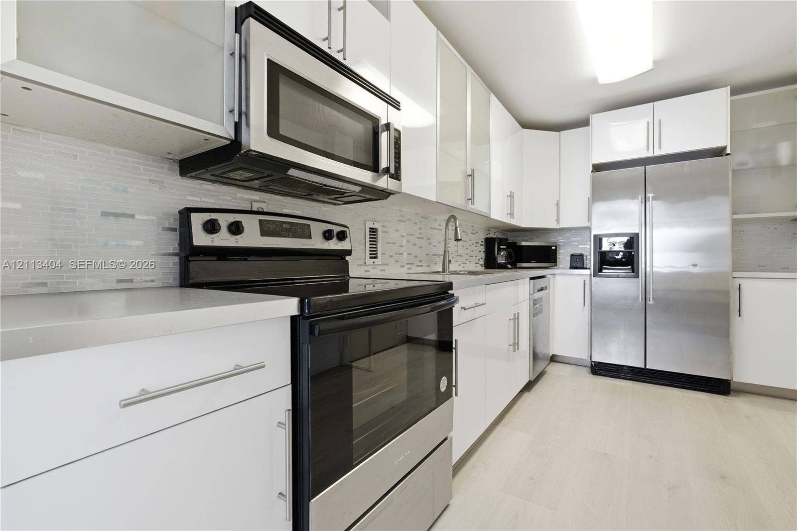 Kitchen featuring high-gloss white cabinetry, stainless steel appliances, light-toned hard-surface flooring, and a linear tile backsplash