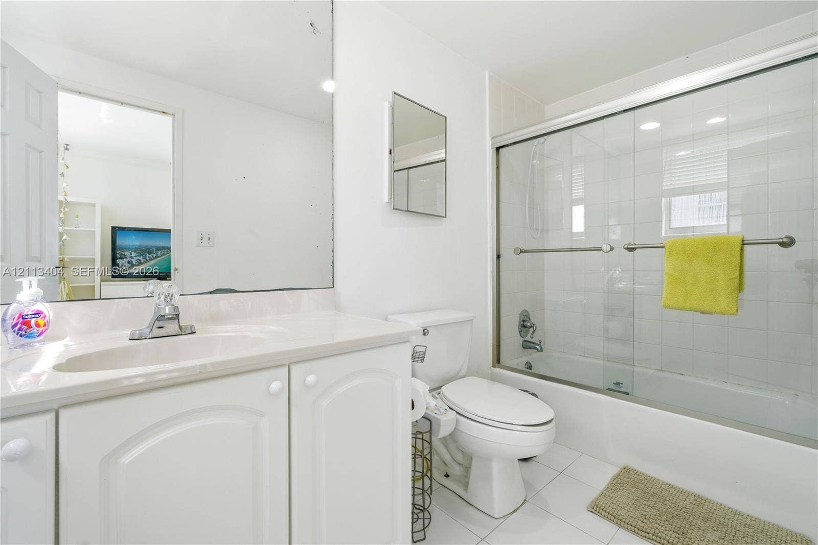 Bathroom featuring a white vanity cabinet, integrated sink with a chrome faucet, large wall-mounted mirror, white ceramic tile flooring, and a bathtub with sliding glass doors and white tile surround