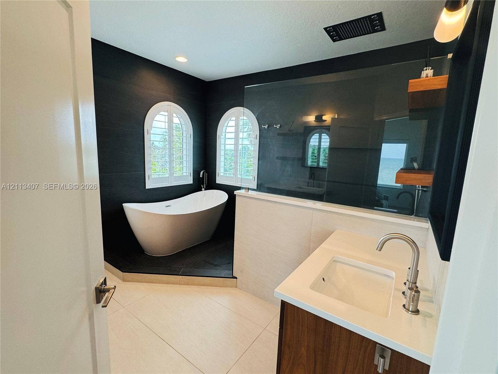 Second floor Bathroom featuring a freestanding soaking tub, arched windows with shutters, dark tile accent walls, a vanity with a white countertop and rectangular sink, and floating wood shelving