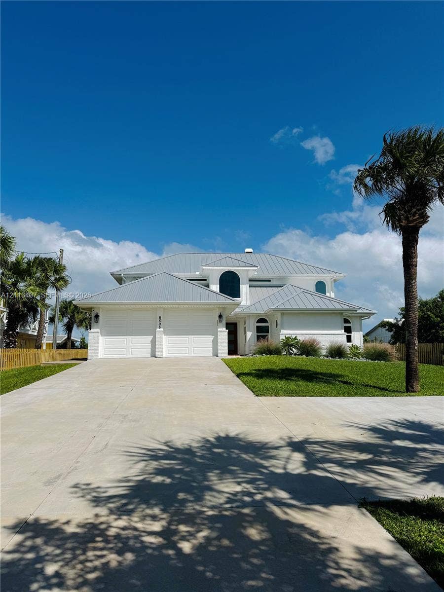 Two-story residence featuring a light-colored stucco exterior, a multi-gabled metal roof, arched window detailing, and an expansive concrete driveway