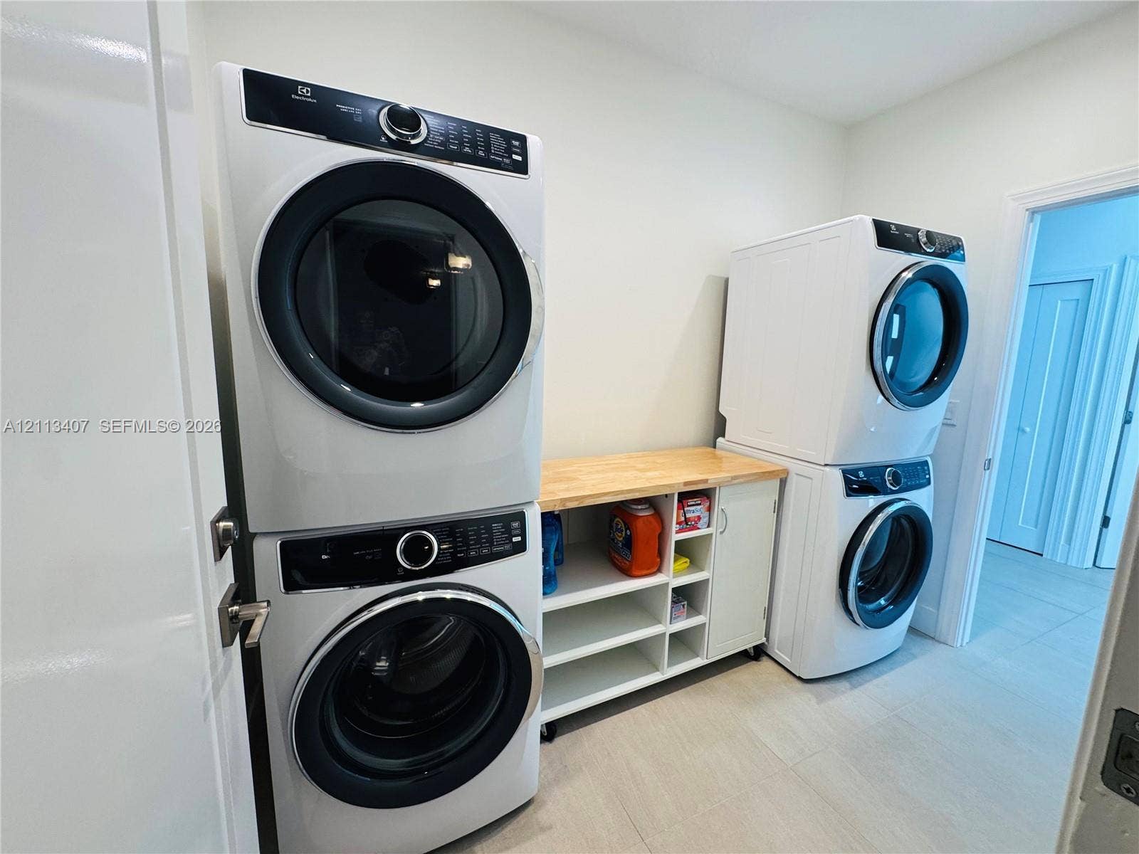 Laundry room featuring dual sets of stackable front-loading washer and dryer units, white custom cabinetry with open shelving, and a natural wood countertop