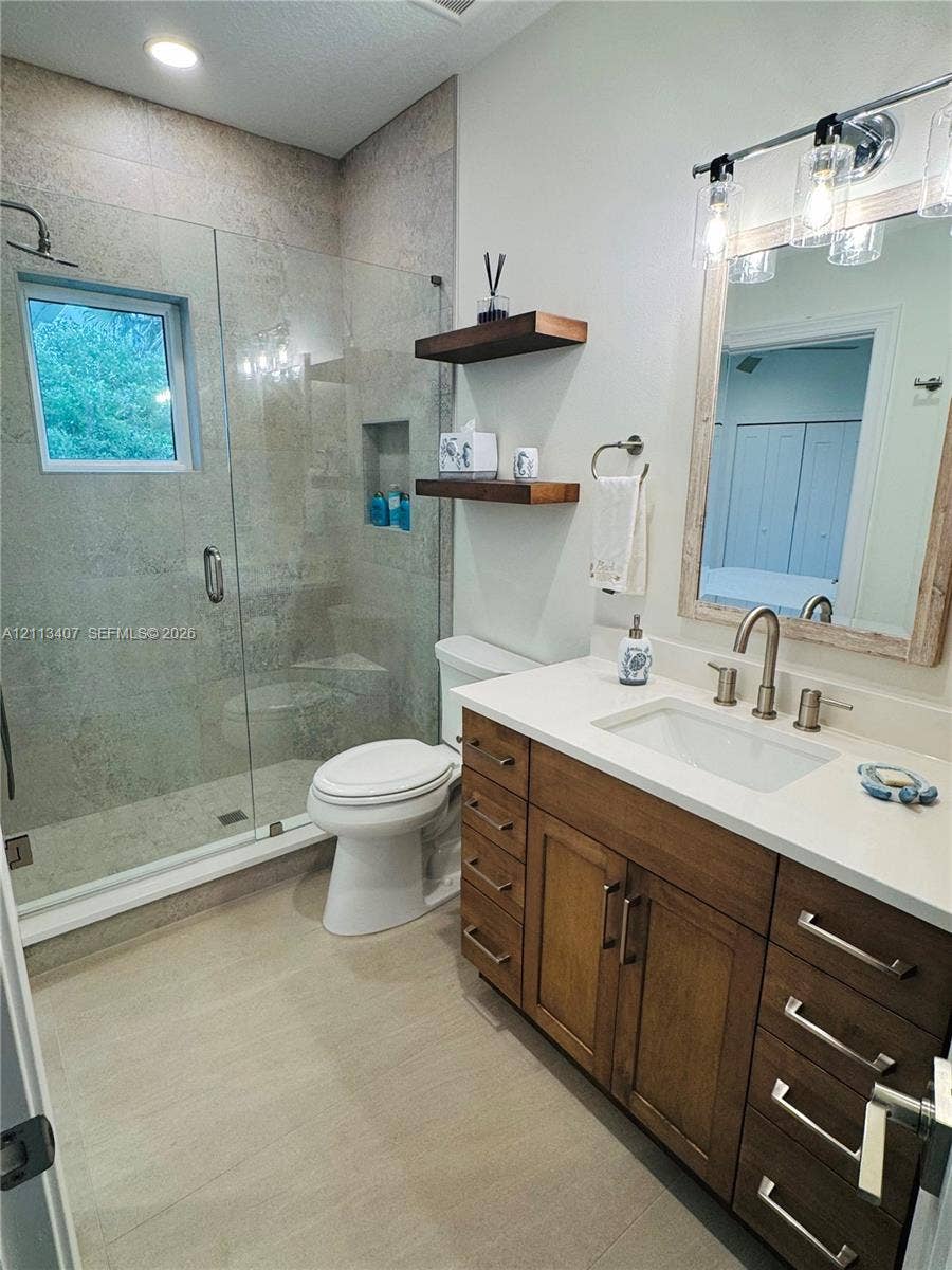 Bathroom featuring a glass-enclosed shower with tiled walls and an integrated niche, wood-finish vanity cabinetry with a light-toned countertop, and two floating wooden shelves