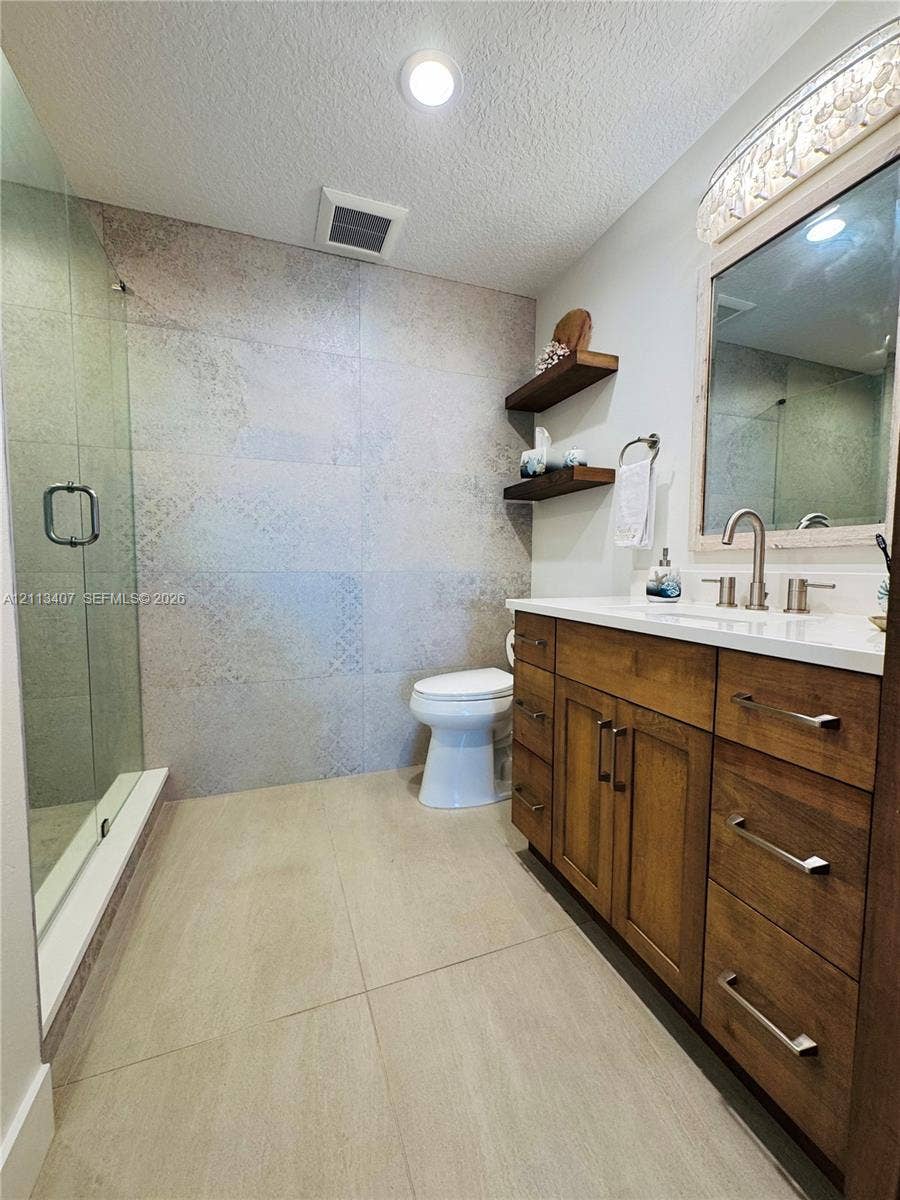 Bathroom featuring a wood-finish vanity with a light-toned countertop, floor-to-ceiling patterned tile shower wall, glass-enclosed shower, dual-tone wall-mounted shelving, and recessed lighting