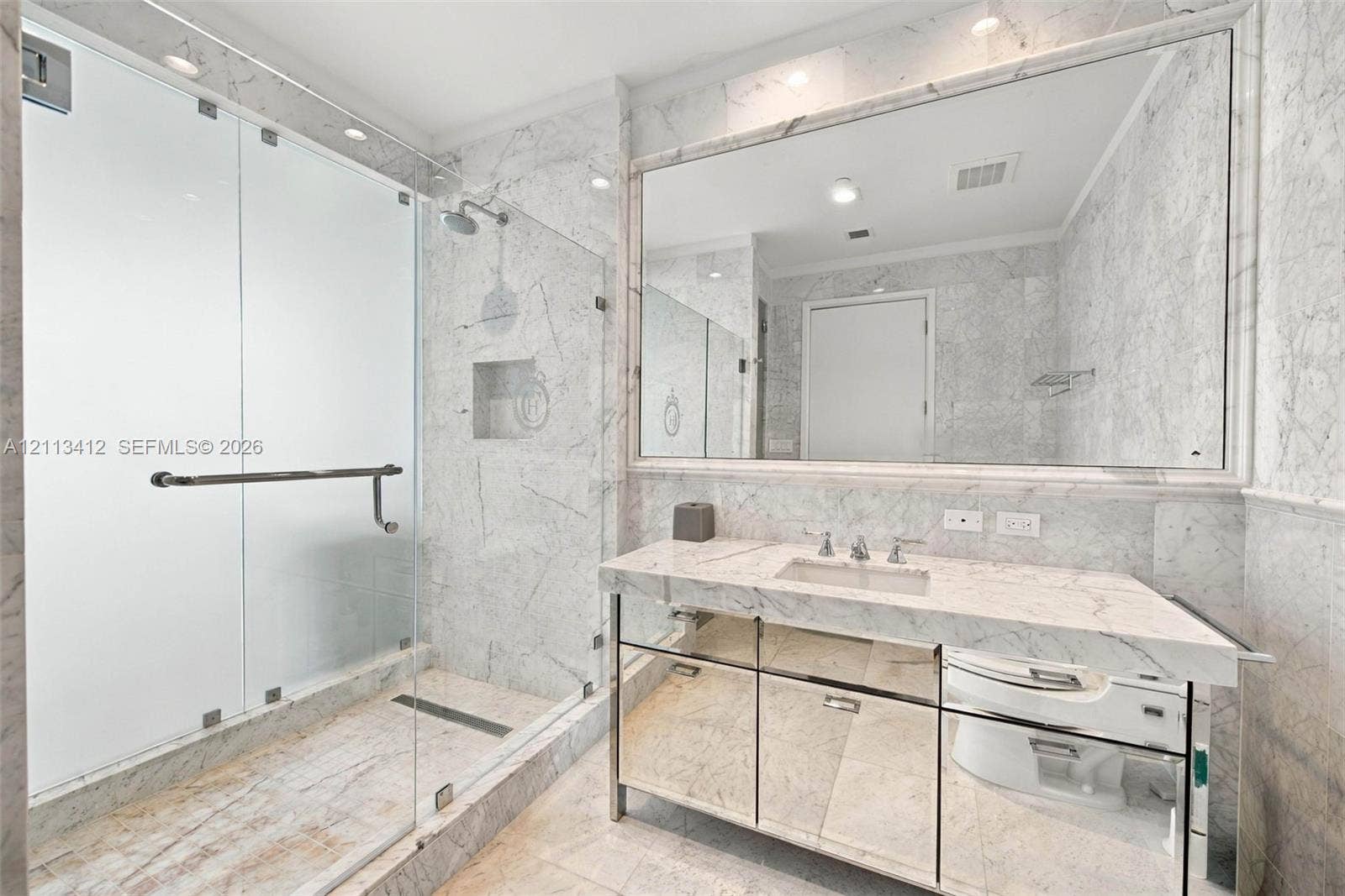 17475 Collins Avenue, #1802, Sunny Isles Beach, FL 33160 Full-height stone wall surfacing, frameless glass shower enclosure, vanity with integrated sink, mirrored cabinet base, and wall-to-wall vanity mirror