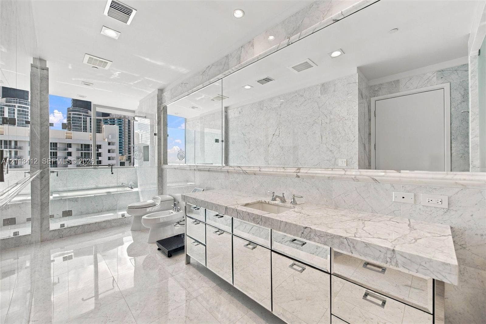 17475 Collins Avenue, #1802, Sunny Isles Beach, FL 33160 Full-length stone vanity with dual undermount sinks, expansive wall-to-wall mirror, floor-to-ceiling natural stone wall and floor tile, glass-enclosed shower, and built-in vanity cabinetry