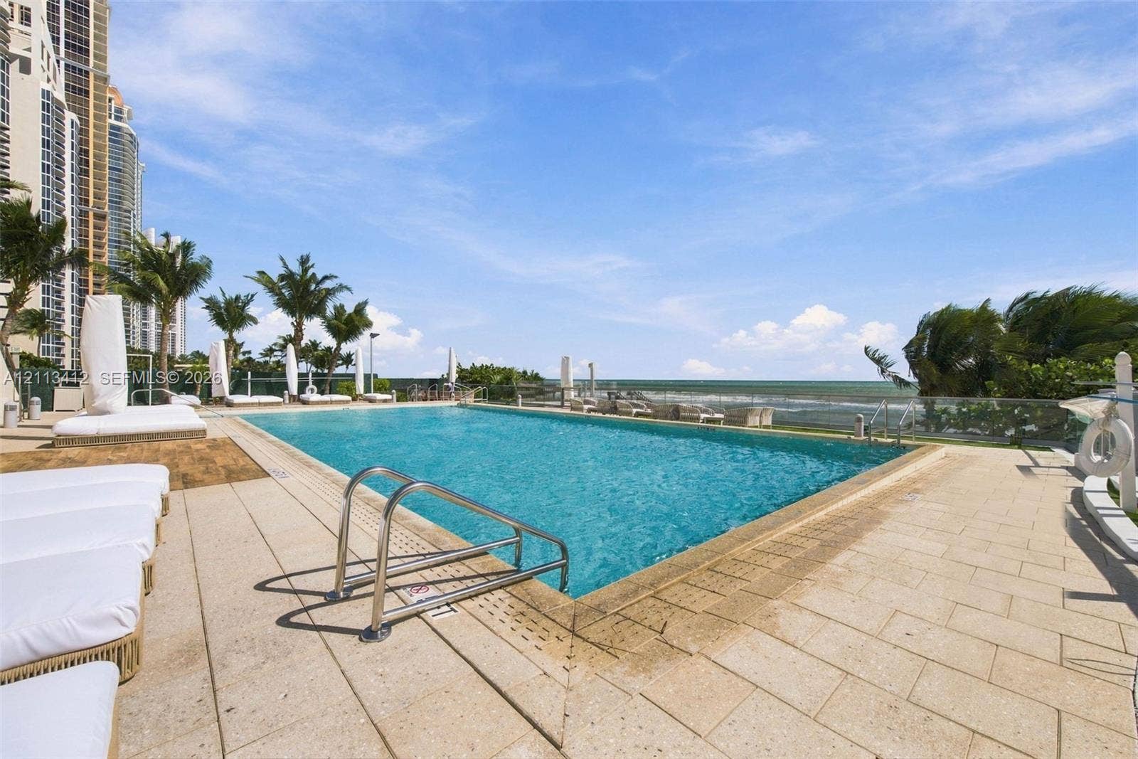 17475 Collins Avenue, #1802, Sunny Isles Beach, FL 33160 Rectangular swimming pool featuring a stone-tile deck, metal entry ladders, glass perimeter fencing, and an ocean horizon