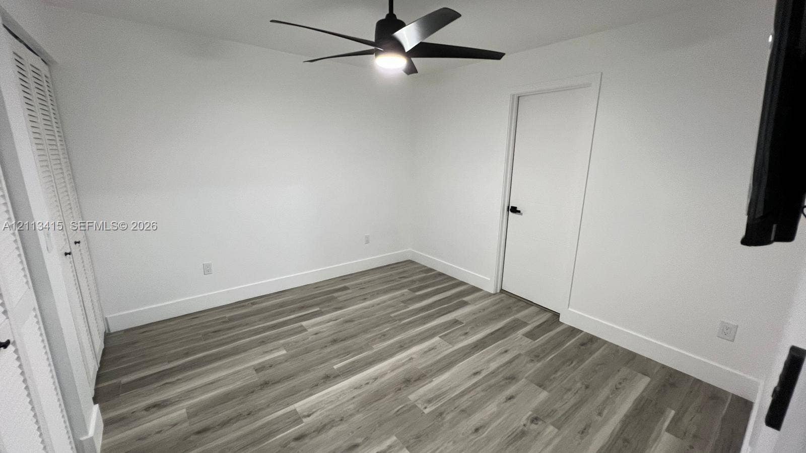 14442 SW 111th Street, #Studio, Miami, Miami, FL 33186 Room featuring wood-finish flooring, white walls, dual louvered-panel closet doors, and a matte black ceiling fan with an integrated light fixture