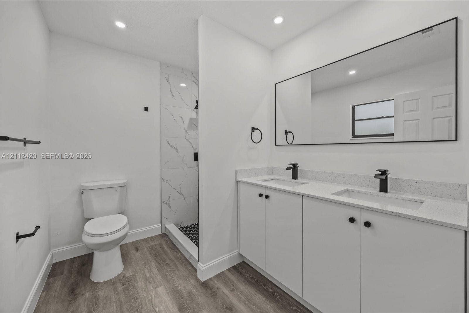Dual-sink vanity with white cabinetry, light-toned stone-finish countertop, walk-in shower with stone-patterned tile, matte black fixtures, and wood-finish flooring