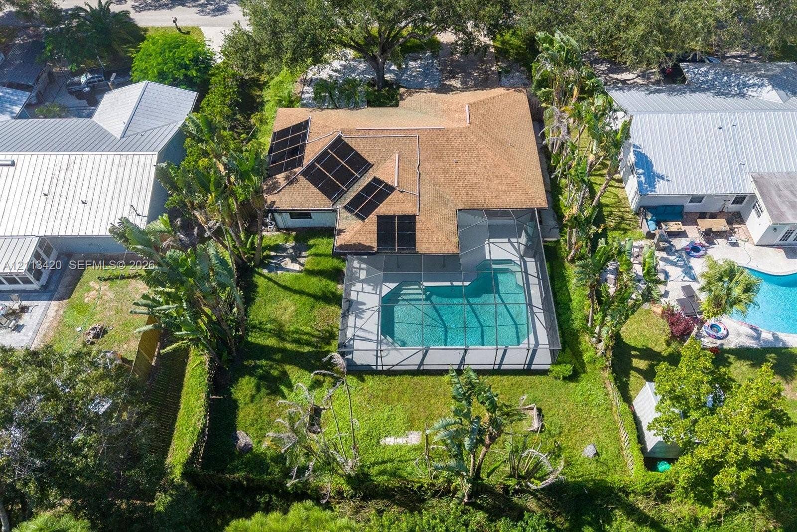 Aerial perspective of a property featuring an in-ground pool enclosed within a screened lanai, roof-mounted solar panels, mature landscaping, and a fenced perimeter