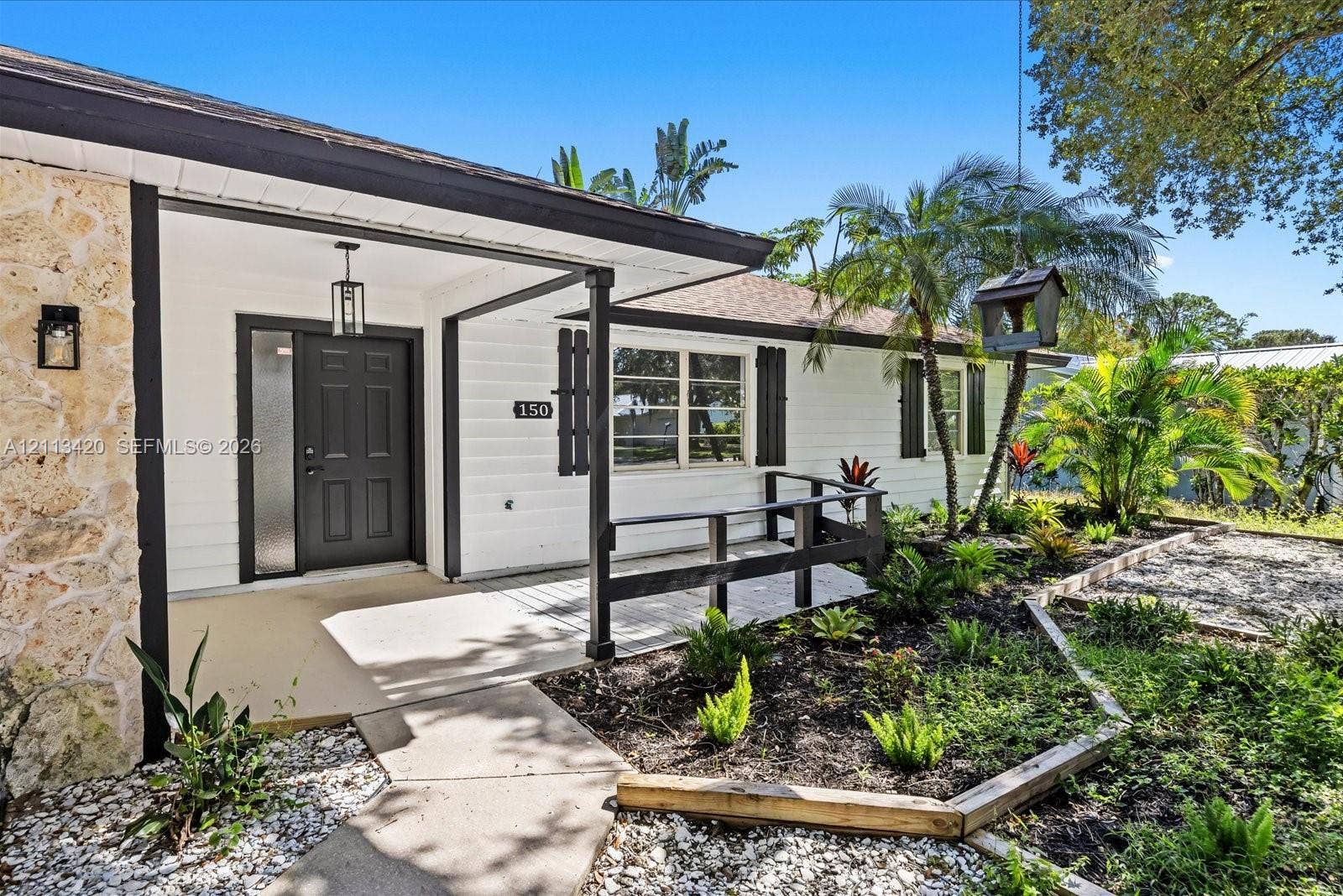 Exterior featuring a stone facade, horizontal lap siding, covered porch with dark-toned railings, and mature tropical landscaping