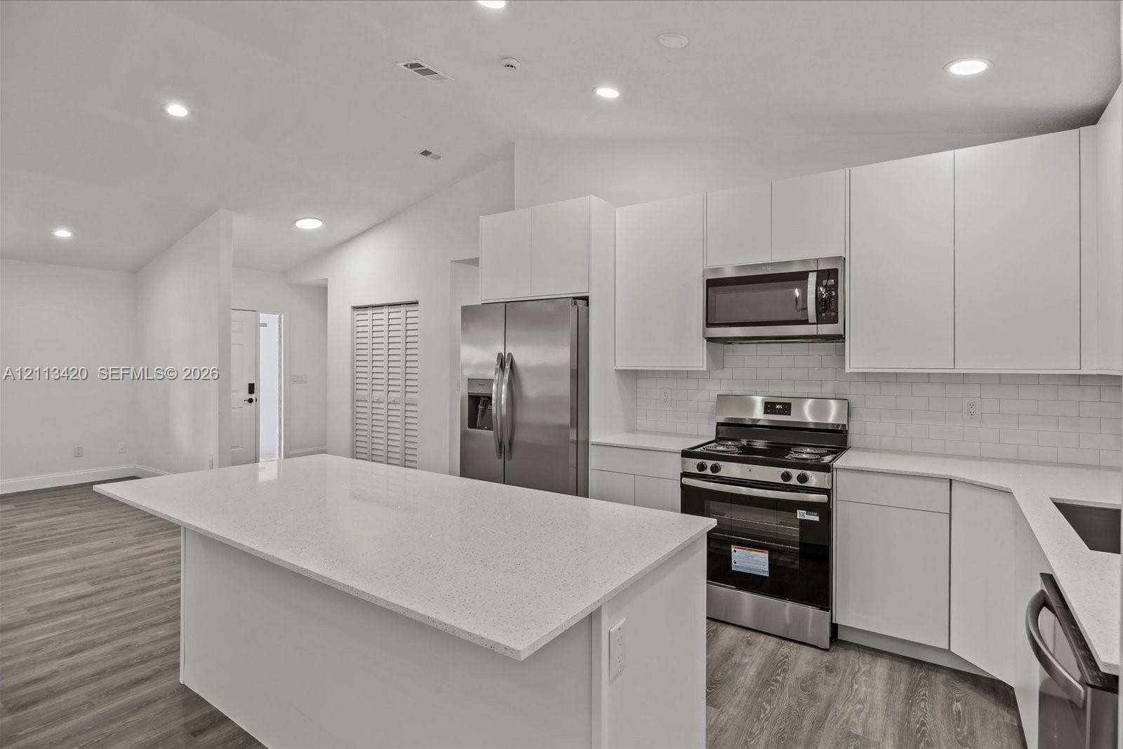 Kitchen featuring white shaker-style cabinetry, an expansive central island with stone-surface countertop, stainless steel appliances, subway tile backsplash, and wood-finish flooring