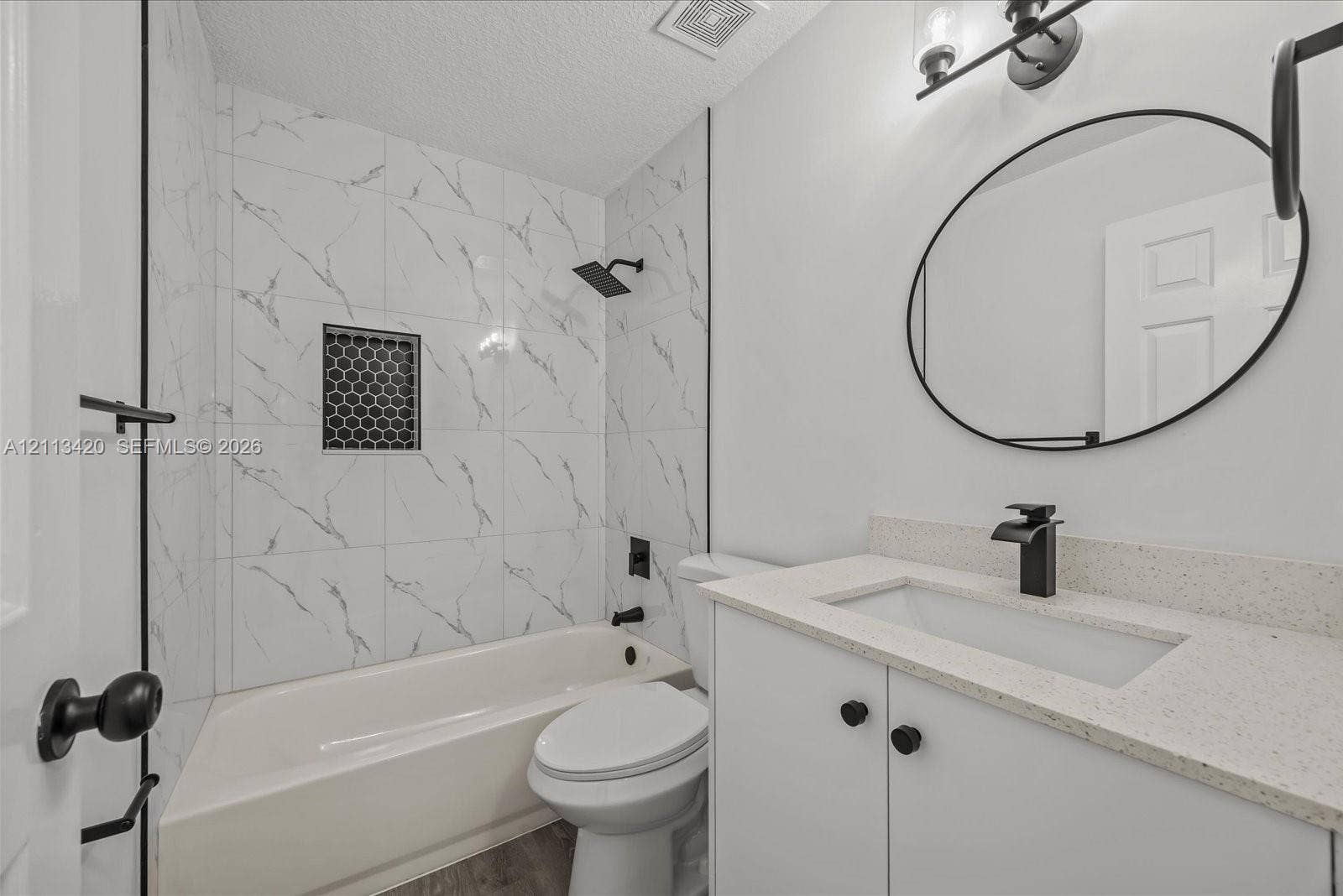 Bathroom featuring stone-finish wall tile in the tub surround with a hexagon-tile niche, matte black rainfall shower fixture, quartz-surround vanity, oval wall-mounted mirror, and matte black faucet hardware