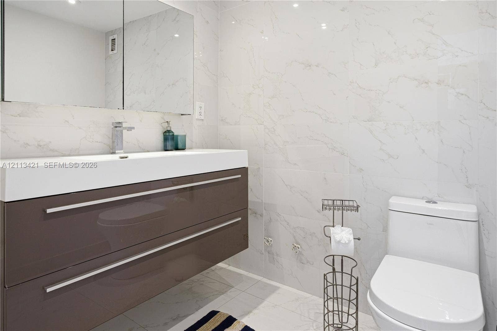 3901 S Ocean Drive, #5L, Hollywood, FL 33019 Floating vanity featuring a high-gloss finish, integrated sink, and polished chrome hardware, paired with floor-to-ceiling stone-patterned tile walls, a mirrored medicine cabinet, and a contemporary one-piece toilet