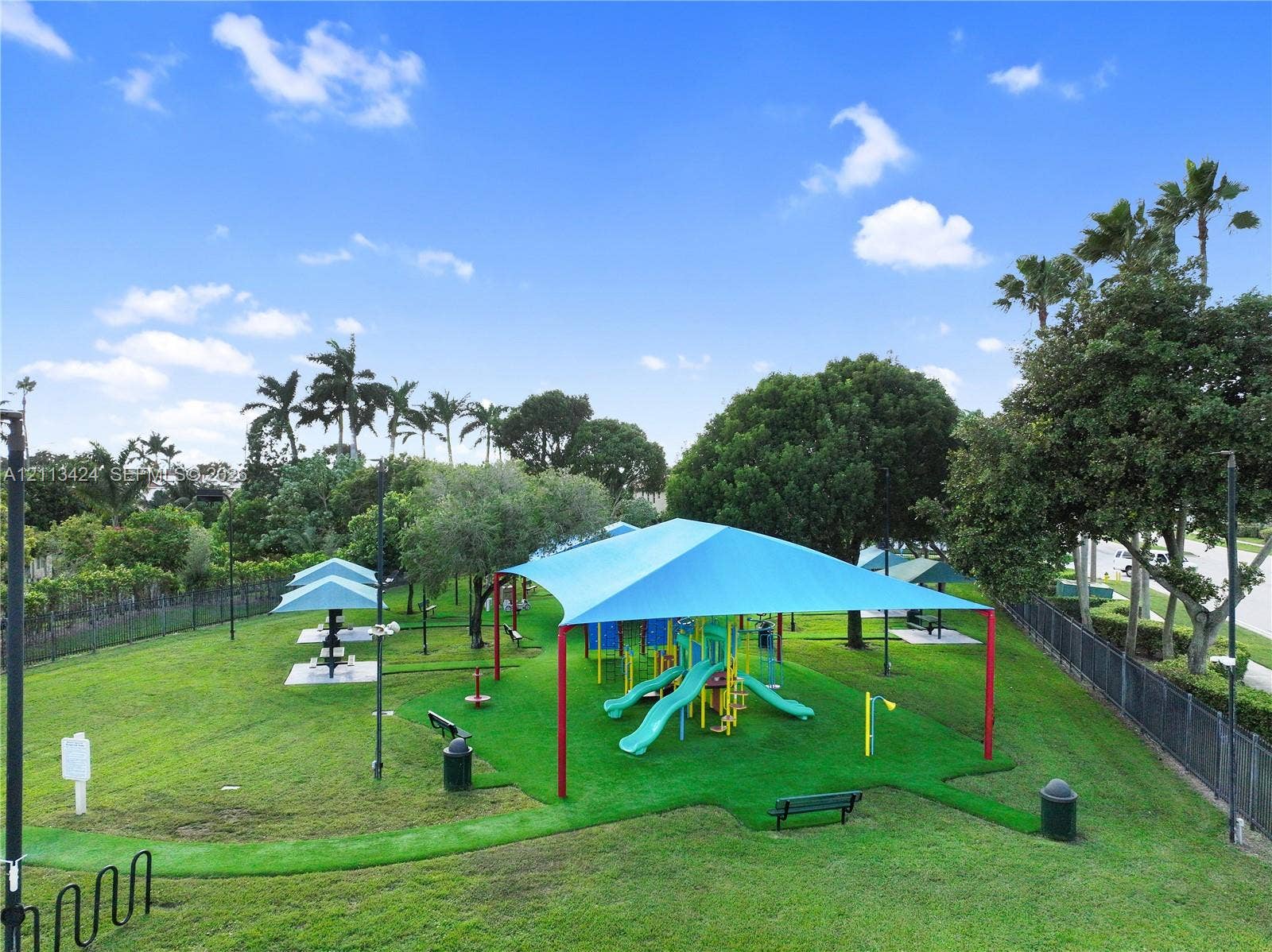 2465 SW 132nd Terrace, Miramar, FL 33027 Outdoor recreation area featuring a playground structure with a blue shade canopy, integrated turf surfacing, picnic stations, perimeter fencing, and mature landscaping