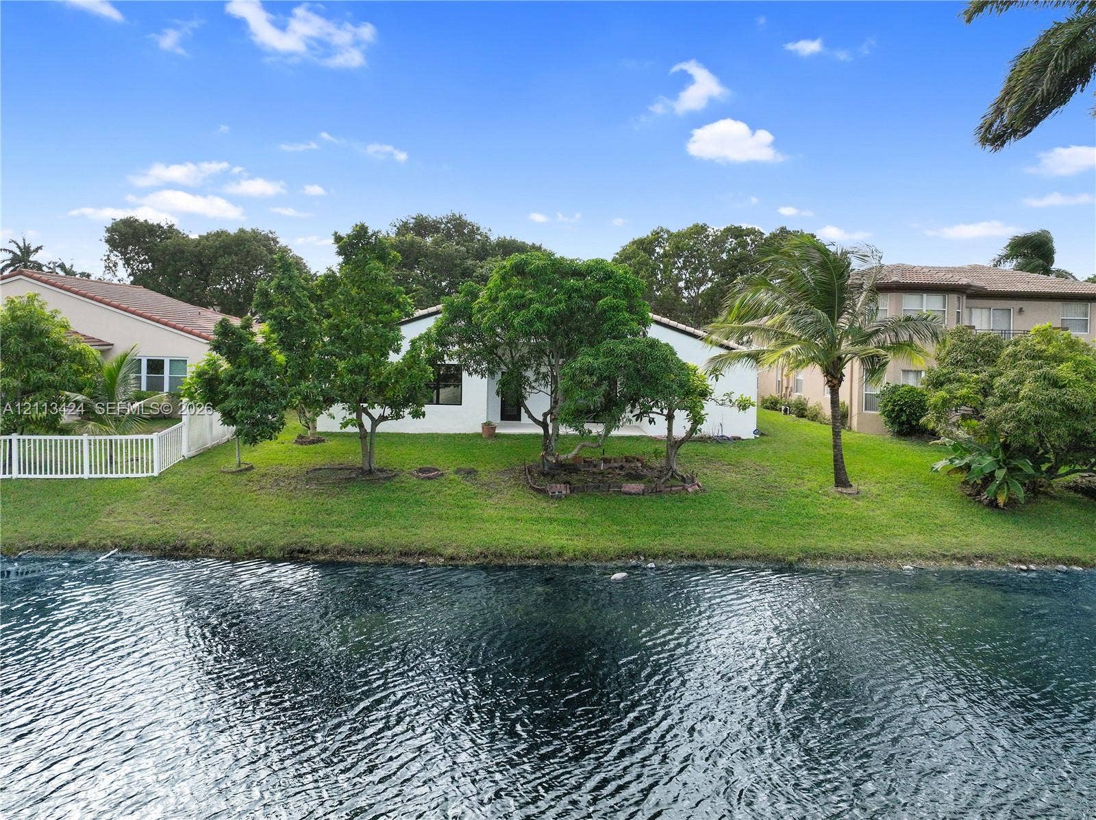 2465 SW 132nd Terrace, Miramar, FL 33027 Waterfront property featuring a manicured lawn, mature trees, and a white privacy fence