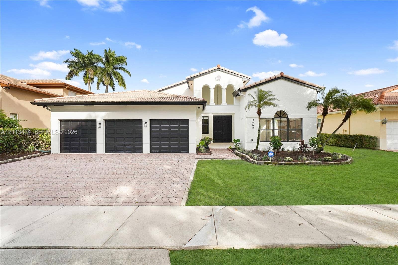 2465 SW 132nd Terrace, Miramar, FL 33027 Stucco exterior featuring a tile roof, three-car garage with paneled doors, arched entryway, and a brick-paver driveway (virtually edited out tree)