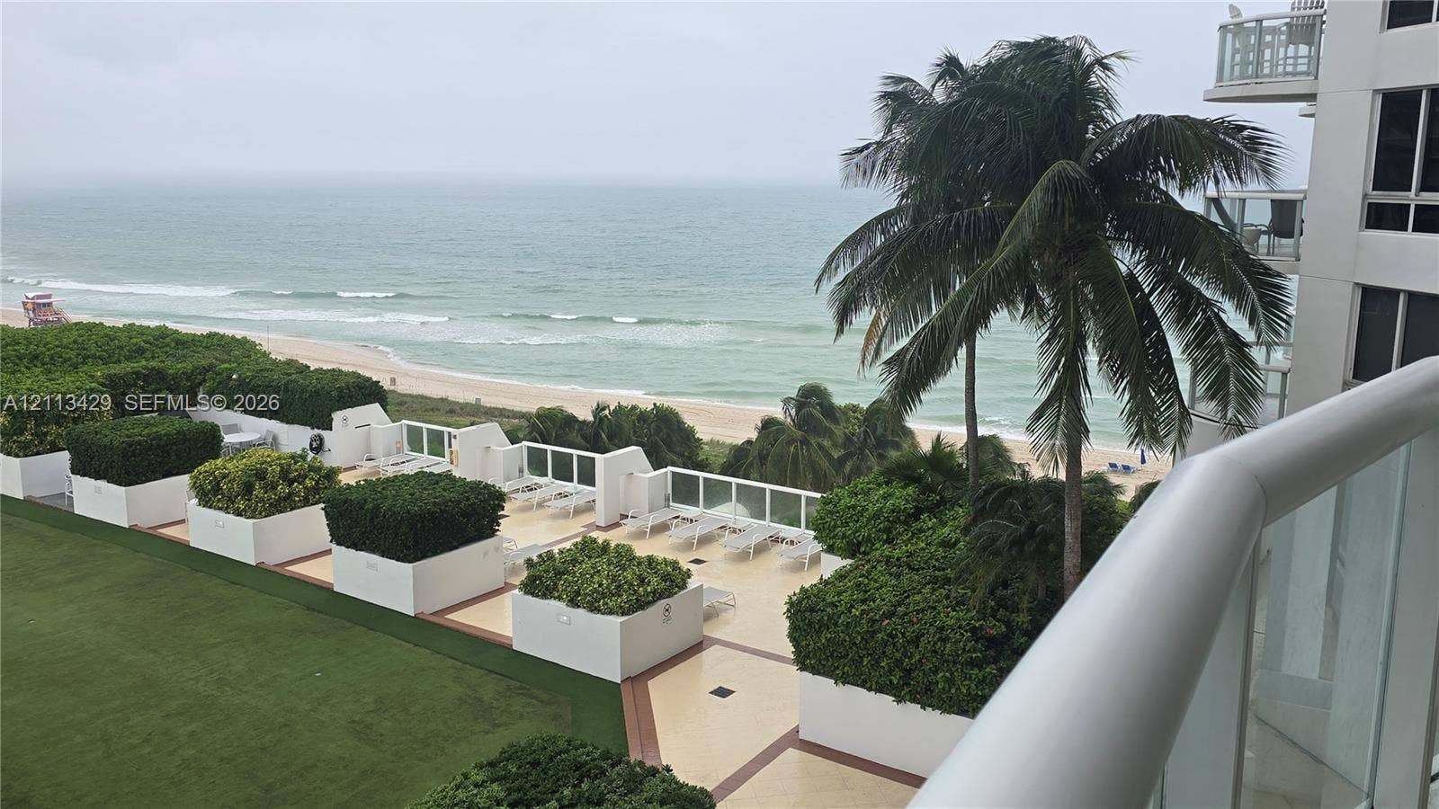 6365 Collins Avenue, #909, Miami Beach, FL 33141 Elevated terrace featuring white glass-panel railings, tile flooring, built-in planter boxes with mature greenery, and direct oceanfront scenery
