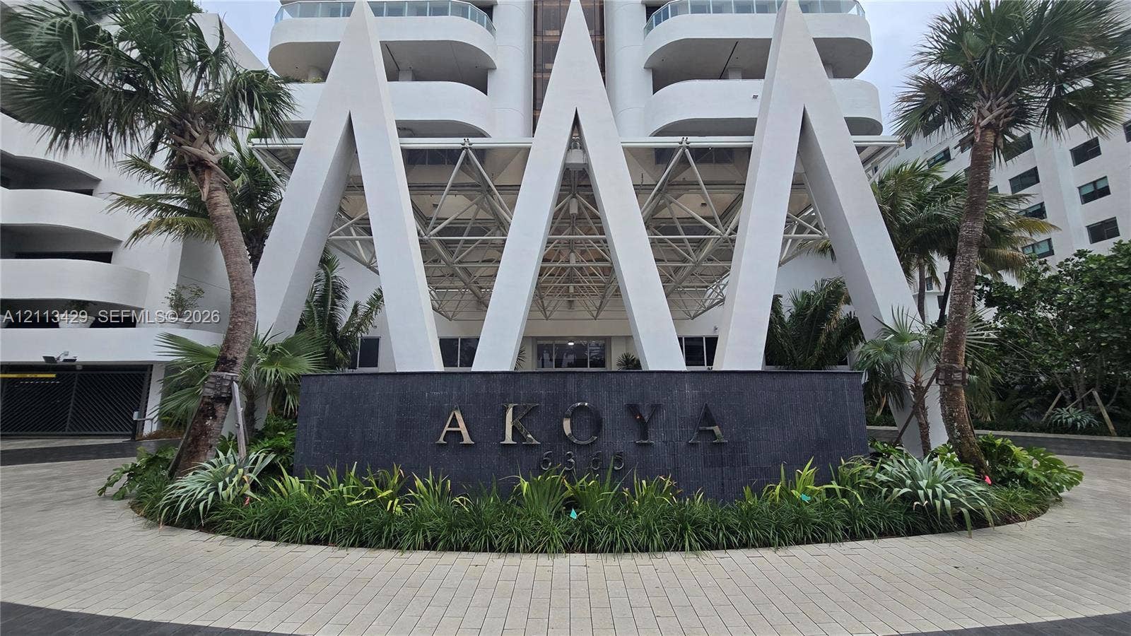 6365 Collins Avenue, #909, Miami Beach, FL 33141 High-rise exterior featuring prominent white geometric structural detailing, a dark stone entrance monument with architectural signage, a paved circular motor court, and mature palm landscaping