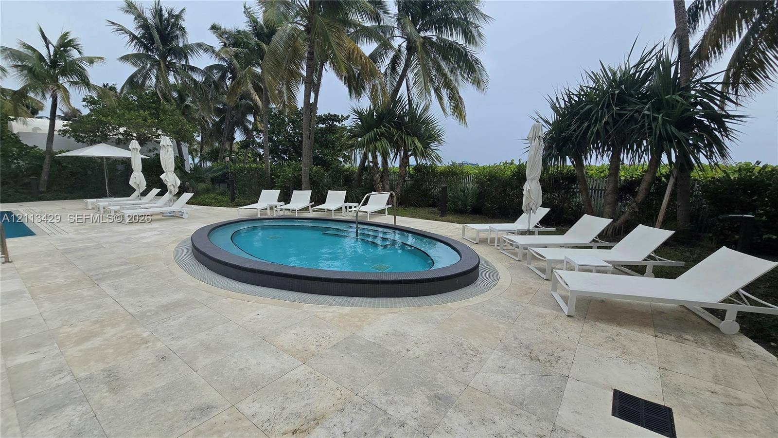 6365 Collins Avenue, #909, Miami Beach, FL 33141 Oval-shaped spa featuring a dark tiled perimeter, surrounded by light-toned natural stone pavers, integrated pool edge, and mature palm trees