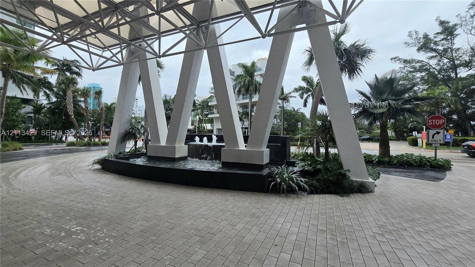 6365 Collins Avenue, #909, Miami Beach, FL 33141 Covered porte-cochre featuring geometric structural supports, stone-paver driveway, and a built-in water feature with lush landscaping