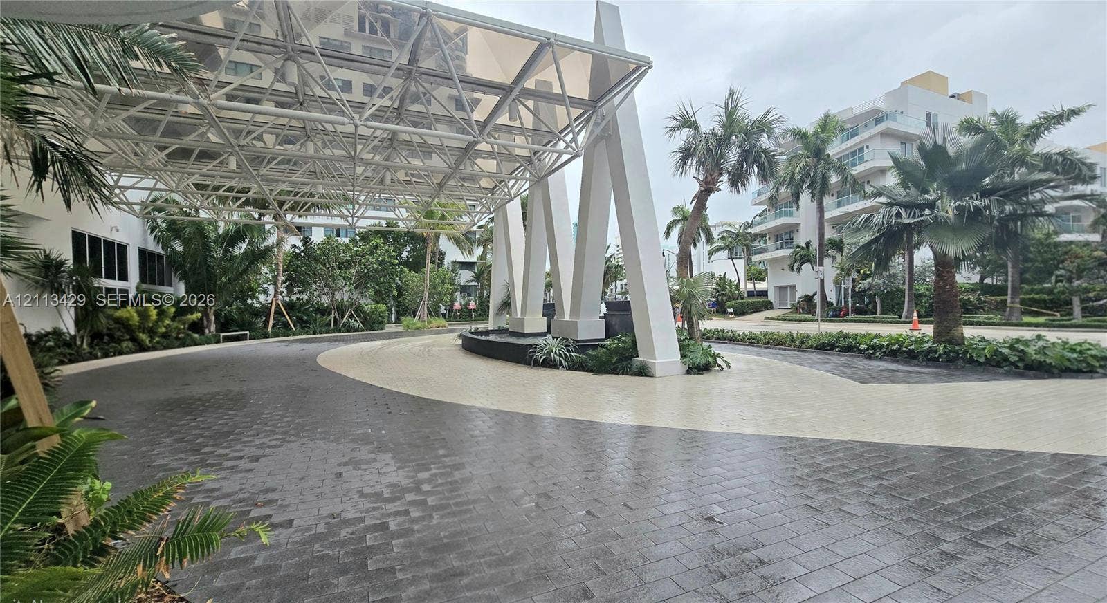 6365 Collins Avenue, #909, Miami Beach, FL 33141 Covered porte-cochre featuring a structural geometric canopy, decorative stone masonry fountain, paved circular driveway, and manicured tropical landscaping