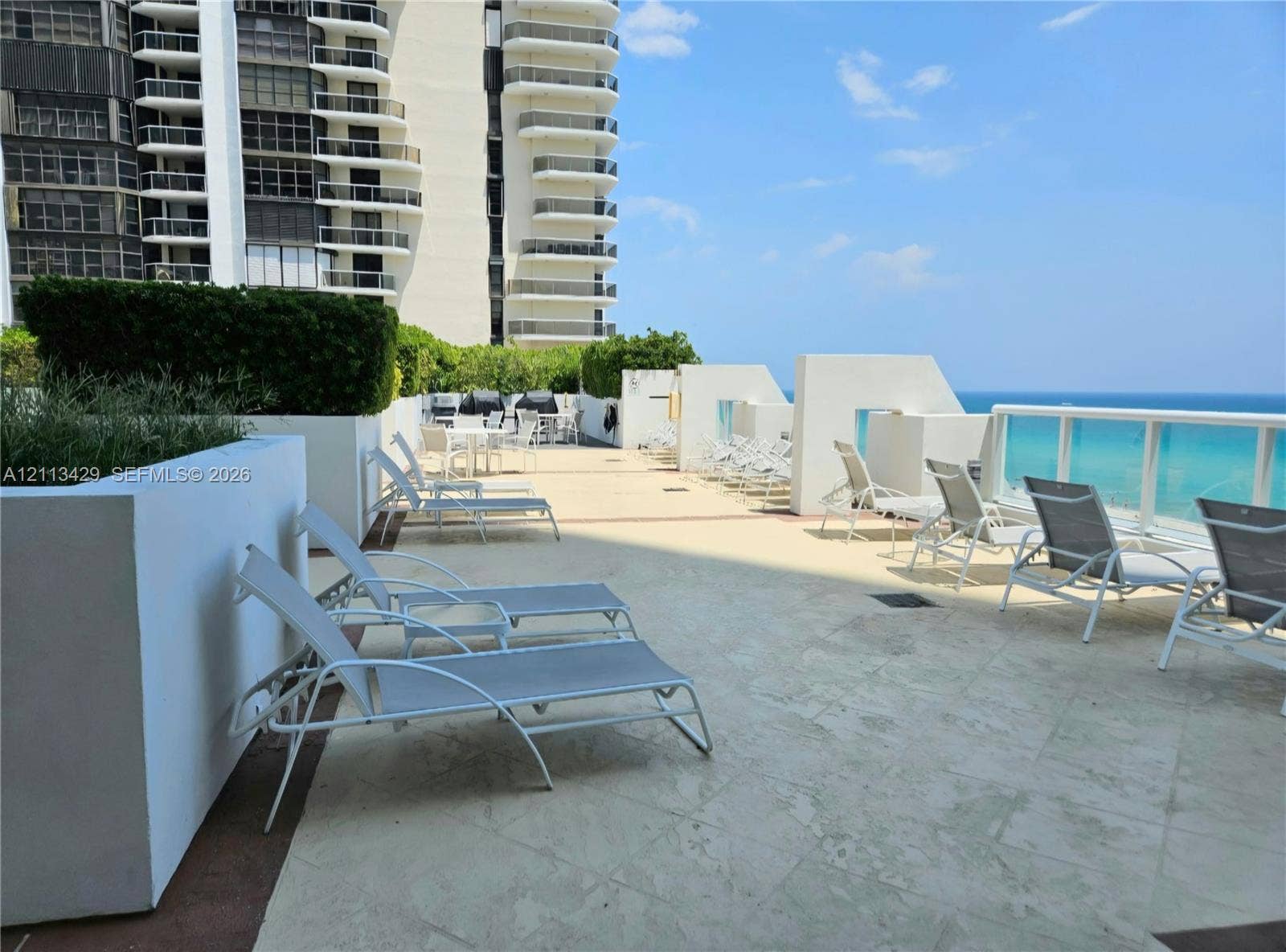 6365 Collins Avenue, #909, Miami Beach, FL 33141 Expansive outdoor terrace featuring stone-tiled flooring, white masonry planters with manicured greenery, and glass-panel railing