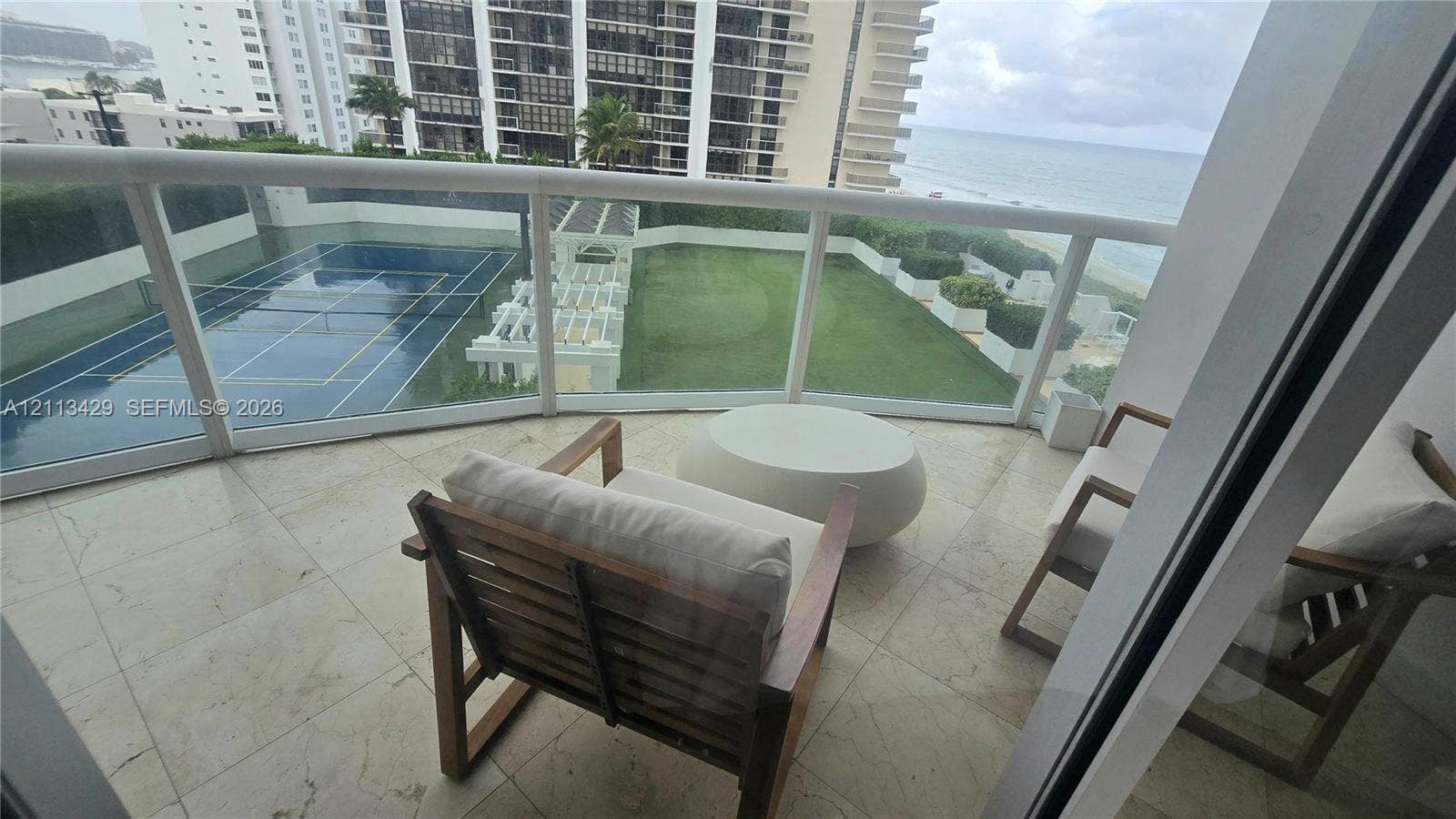 6365 Collins Avenue, #909, Miami Beach, FL 33141 Balcony featuring stone tile flooring and a glass-paneled railing, overlooking a hard-surfaced tennis court, a manicured lawn, and a white architectural pergola