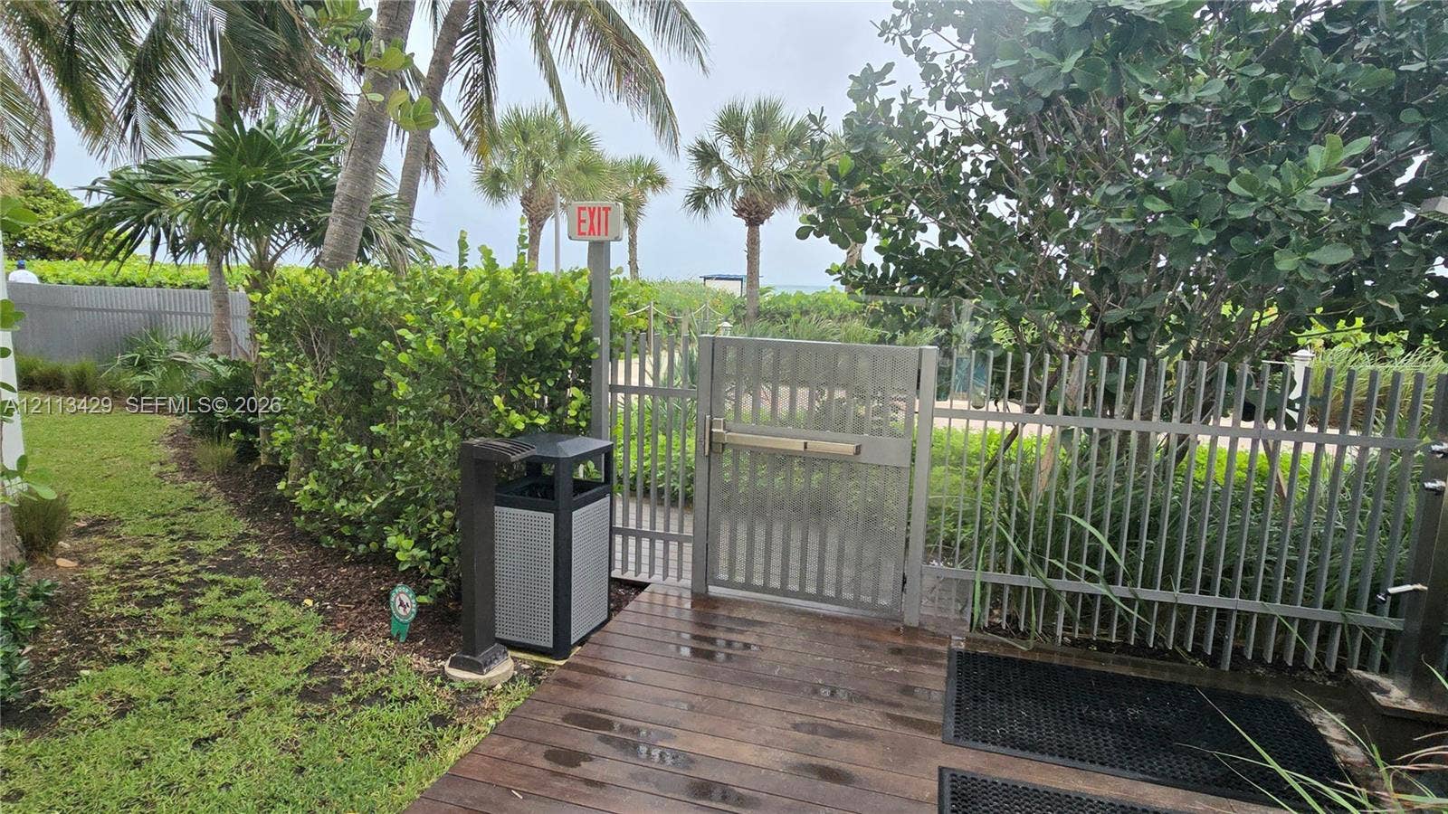 6365 Collins Avenue, #909, Miami Beach, FL 33141 Outdoor area featuring a wood-finish deck, metal perimeter fencing with a gated entry, manicured landscaping, and mature palm trees