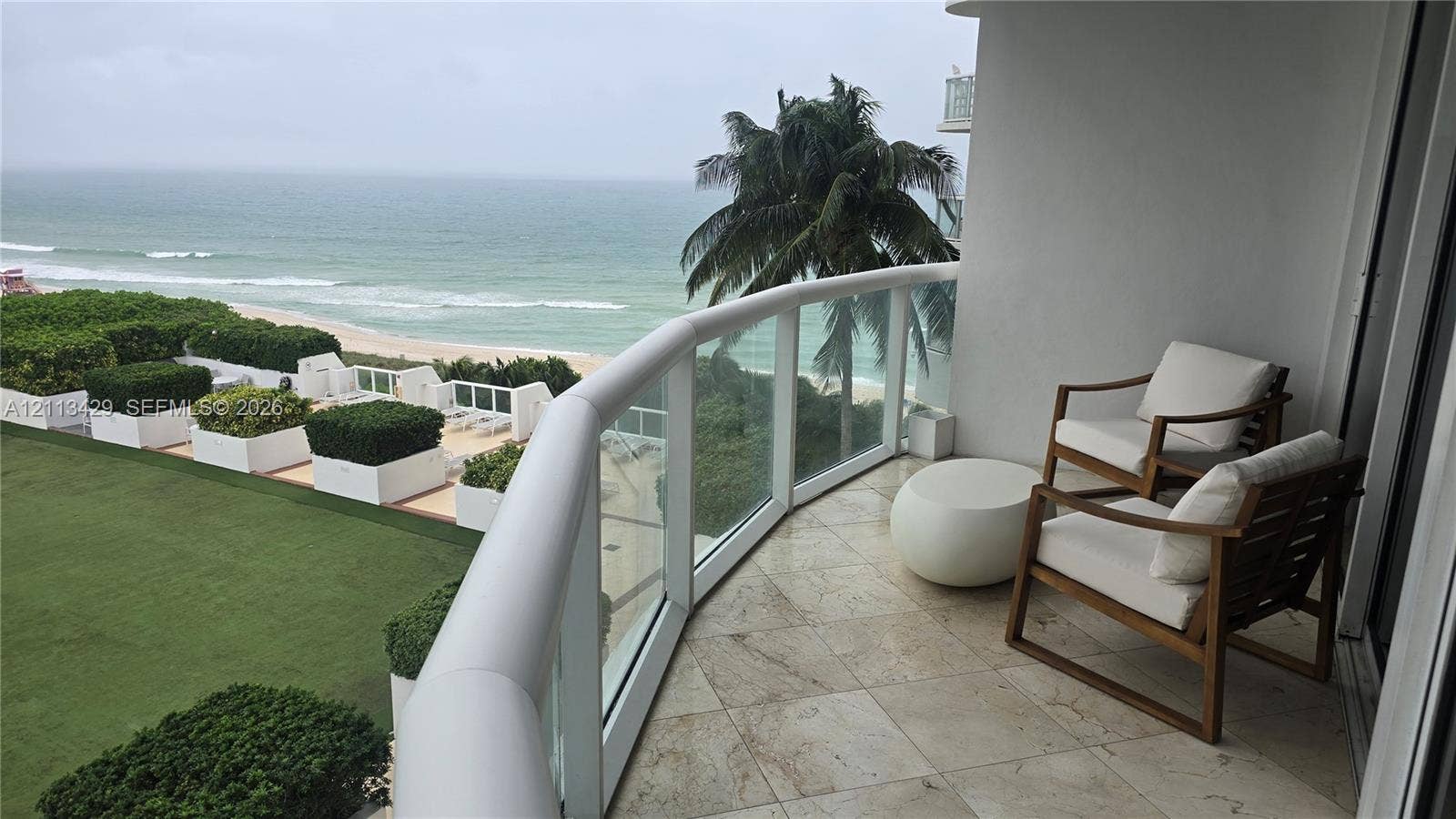 6365 Collins Avenue, #909, Miami Beach, FL 33141 Covered balcony featuring stone tile flooring and a glass panel railing overlooking the oceanfront and manicured grounds