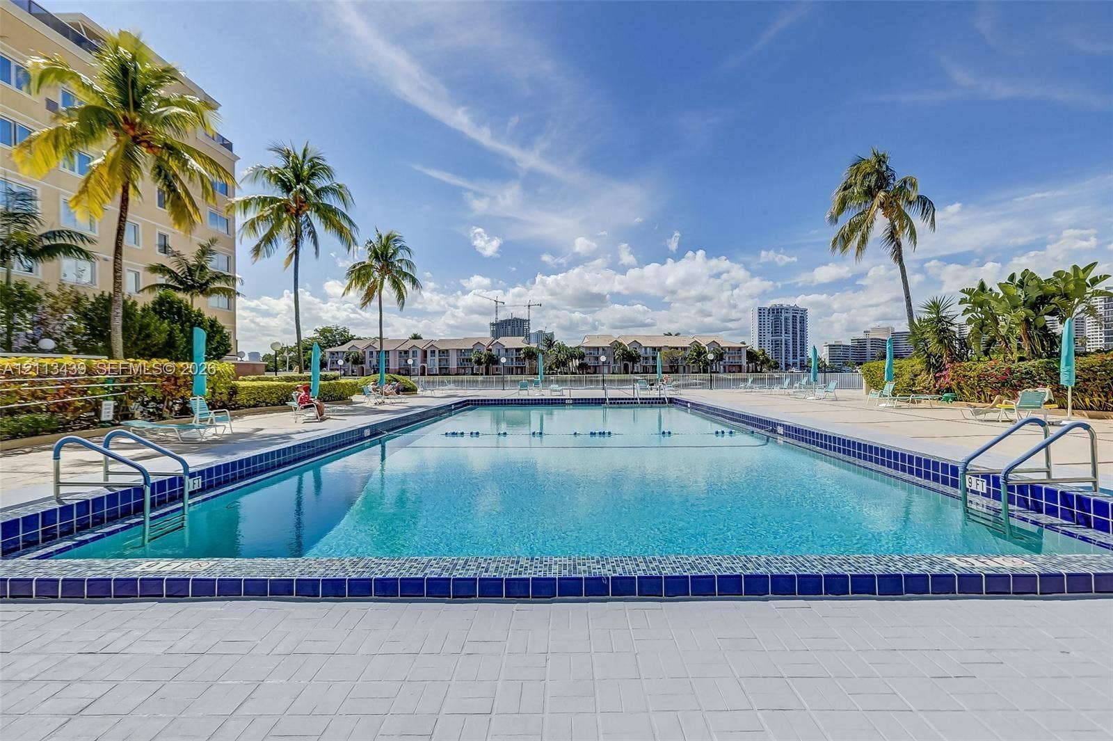 3800 S Ocean Drive, #416, Hollywood, FL 33019 Rectangular swimming pool featuring blue ceramic tile trim, metal entry railings, and an expansive paved pool deck surrounded by palm trees and landscaped hedging