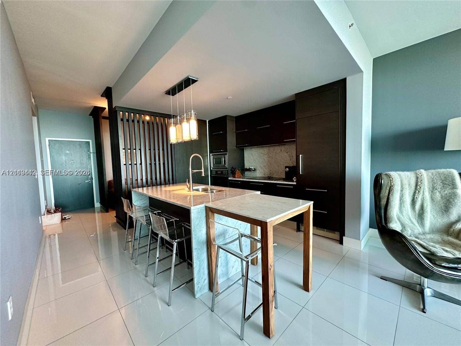900 Biscayne Boulevard, #1903, Miami, Miami, FL 33132 Modern kitchen featuring dark wood cabinetry, a stone-topped island with a waterfall edge, stone backsplash, built-in appliances, and a decorative vertical slat room divider