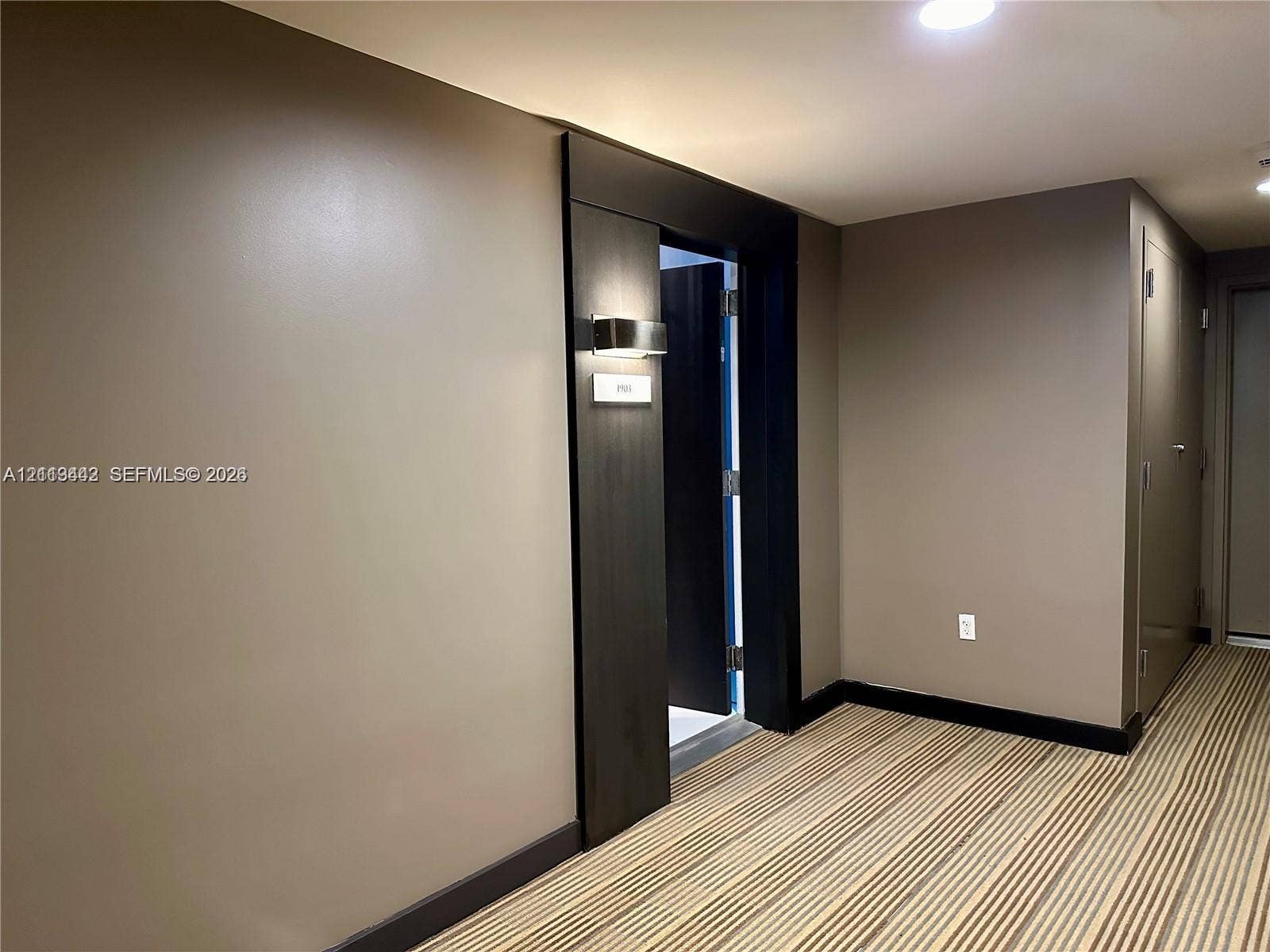 900 Biscayne Boulevard, #1903, Miami, Miami, FL 33132 Neutral-toned interior featuring dark wood-finish trim, a recessed doorway with a wall-mounted metallic sconce, and striped carpeting