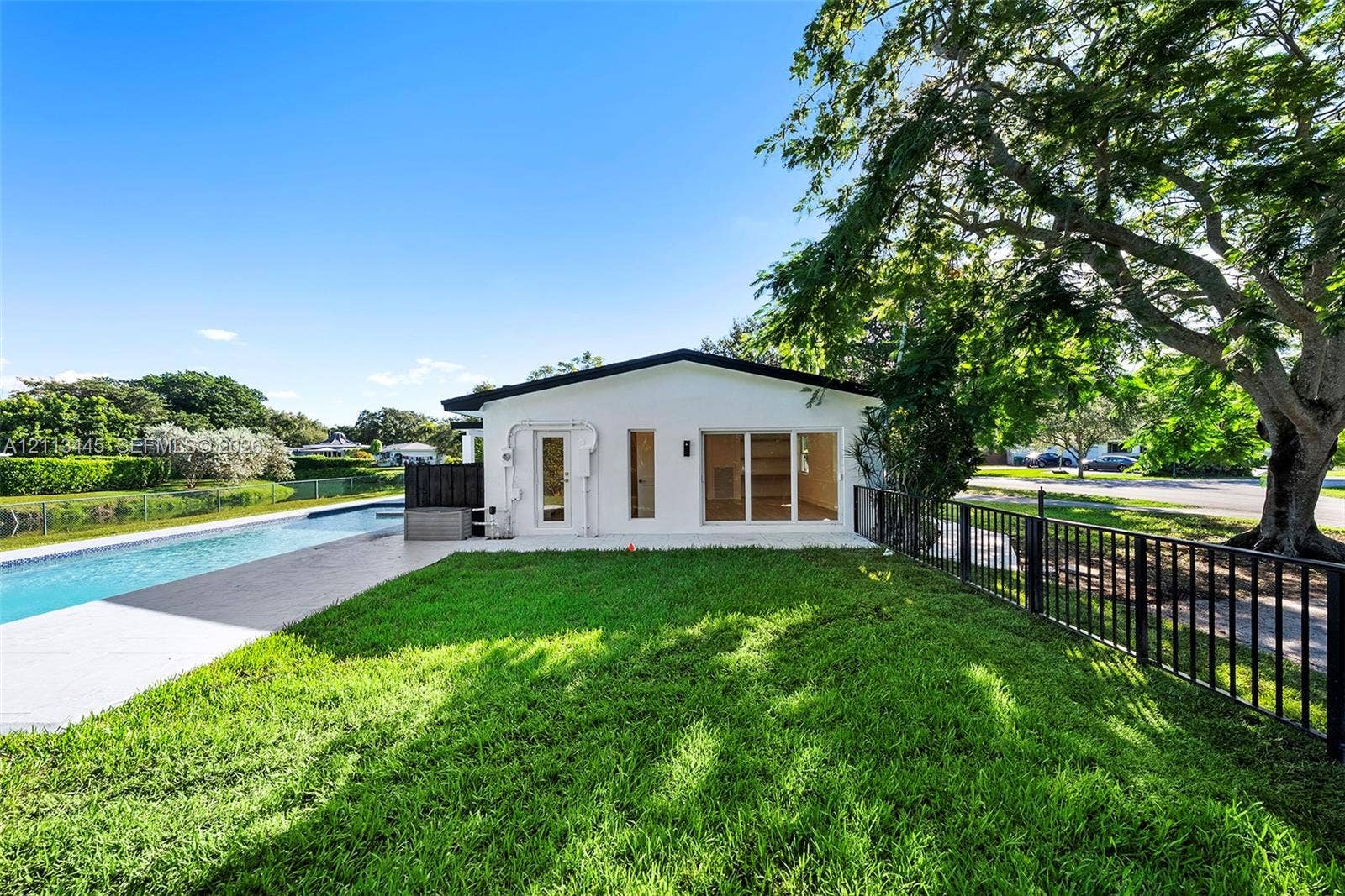 6951 SW 134th Street, Pinecrest, Pinecrest, FL 33156 Exterior rear elevation featuring white stucco siding, a rectangular swimming pool with paved surround, expansive manicured lawn, and black metal perimeter fencing