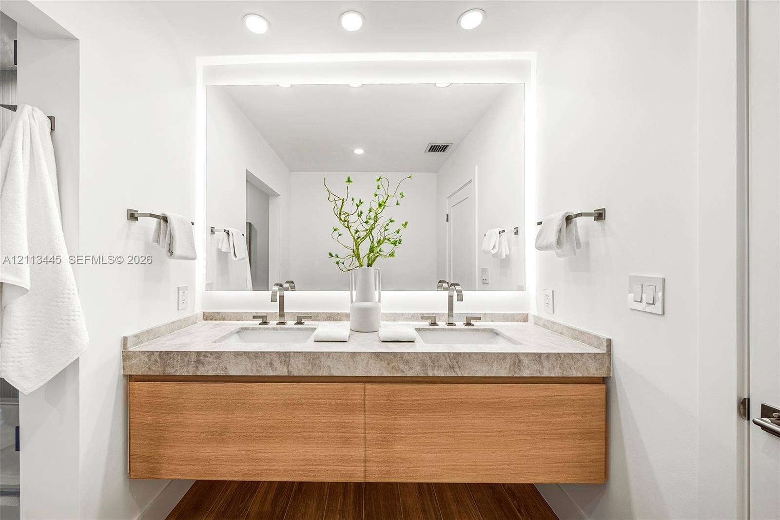 6951 SW 134th Street, Pinecrest, Pinecrest, FL 33156 Floating wood-finish vanity with dual integrated sinks, stone-surface countertop, LED-backlit mirror, and wall-mounted metal fixtures