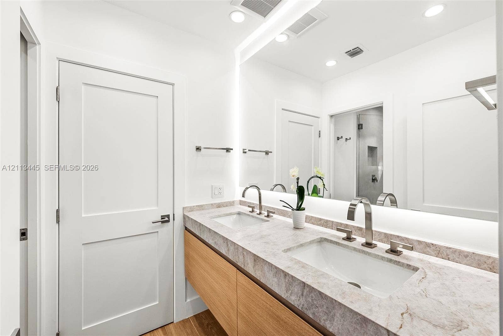6951 SW 134th Street, Pinecrest, Pinecrest, FL 33156 Floating vanity with wood-finish cabinetry, stone countertop, integrated dual undermount sinks, tall arch-neck faucets, and large wall-to-wall mirror with integrated lighting