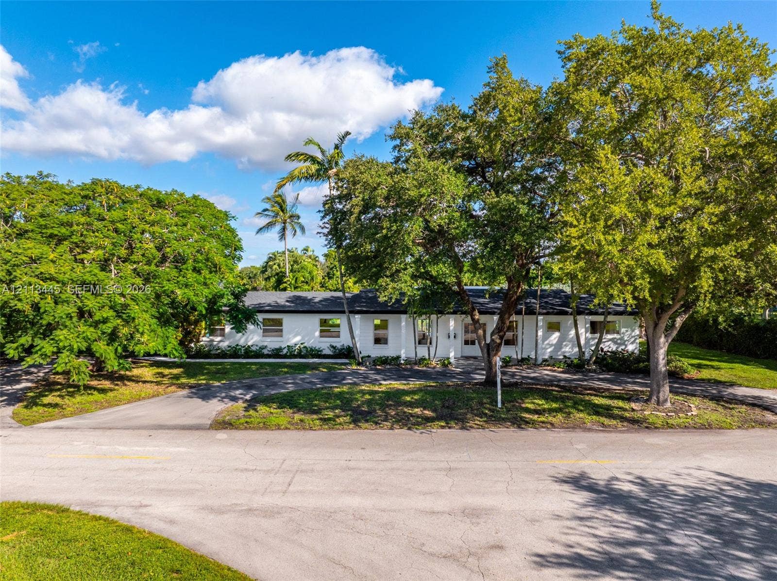 6951 SW 134th Street, Pinecrest, Pinecrest, FL 33156 Single-story exterior featuring white masonry facade, dark shingle roof, paved circular driveway, and mature landscaping