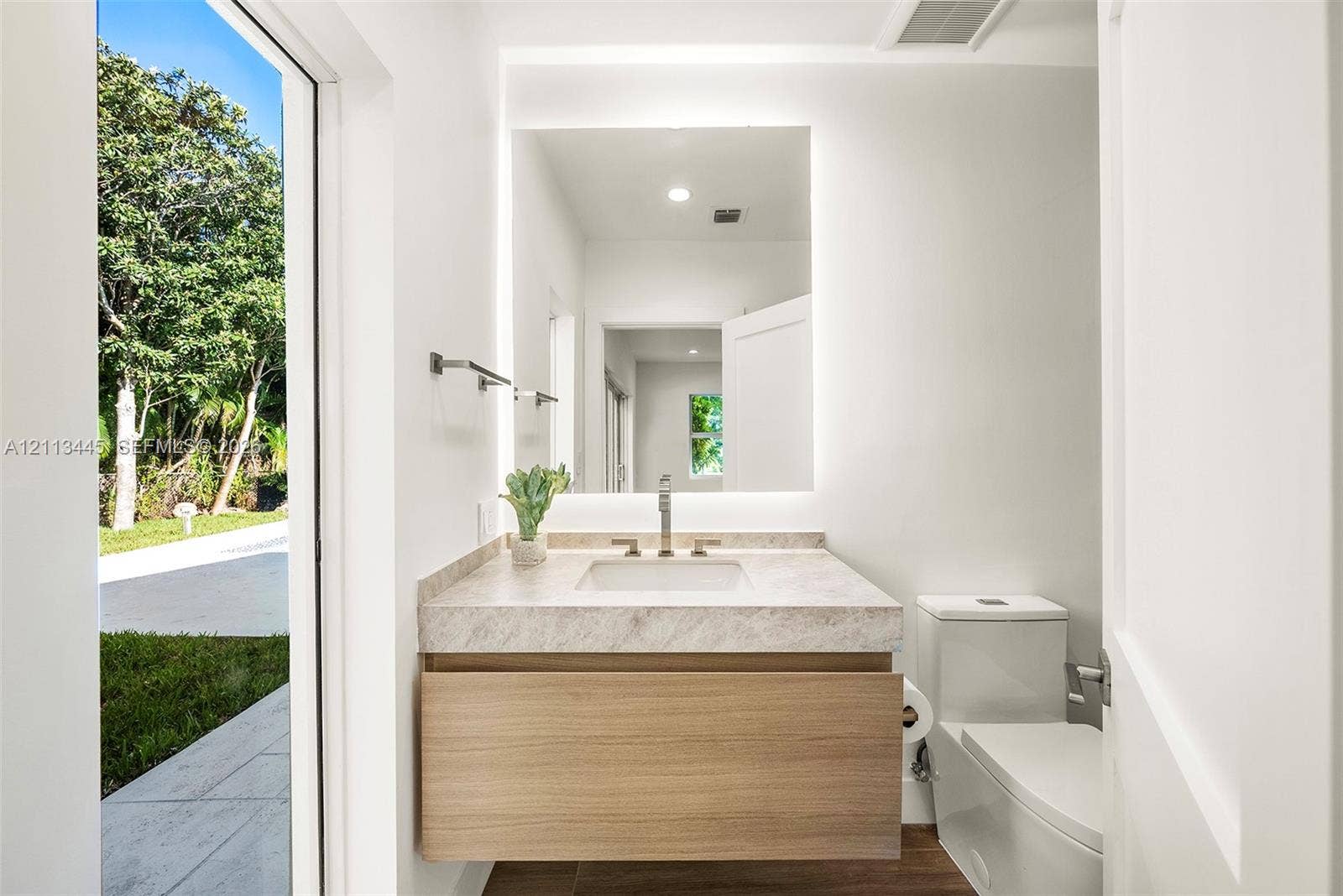 6951 SW 134th Street, Pinecrest, Pinecrest, FL 33156 Floating wood-finish vanity with a stone countertop, rectangular undermount sink, integrated backlit mirror, and dual-flush toilet