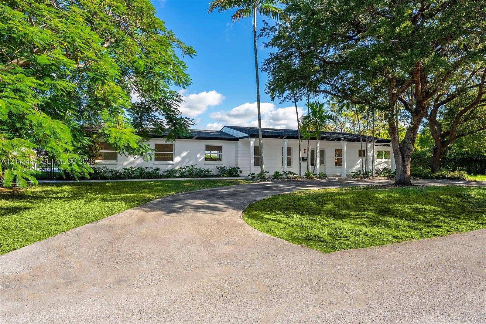 6951 SW 134th Street, Pinecrest, Pinecrest, FL 33156 Single-story exterior featuring white masonry walls, a covered front entryway with structural columns, and an expansive paved driveway set within a landscape of mature trees and established greenery