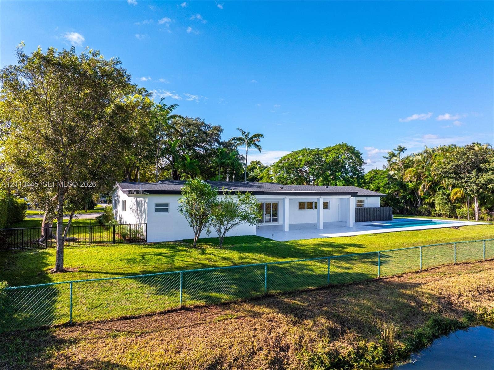 6951 SW 134th Street, Pinecrest, Pinecrest, FL 33156 Exterior featuring a white stucco facade, covered patio, attached rectangular swimming pool, metal perimeter fencing, and mature landscaping