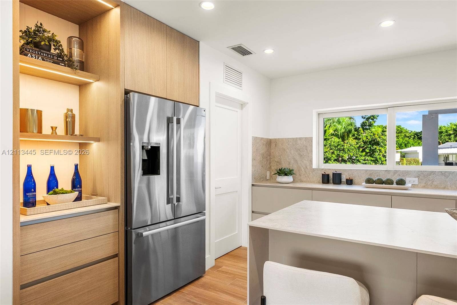 6951 SW 134th Street, Pinecrest, Pinecrest, FL 33156 Kitchen featuring flat-panel cabinetry, light-toned stone-finish countertops, stainless steel refrigerator, integrated wood-finish shelving with recessed lighting, and large horizontal window