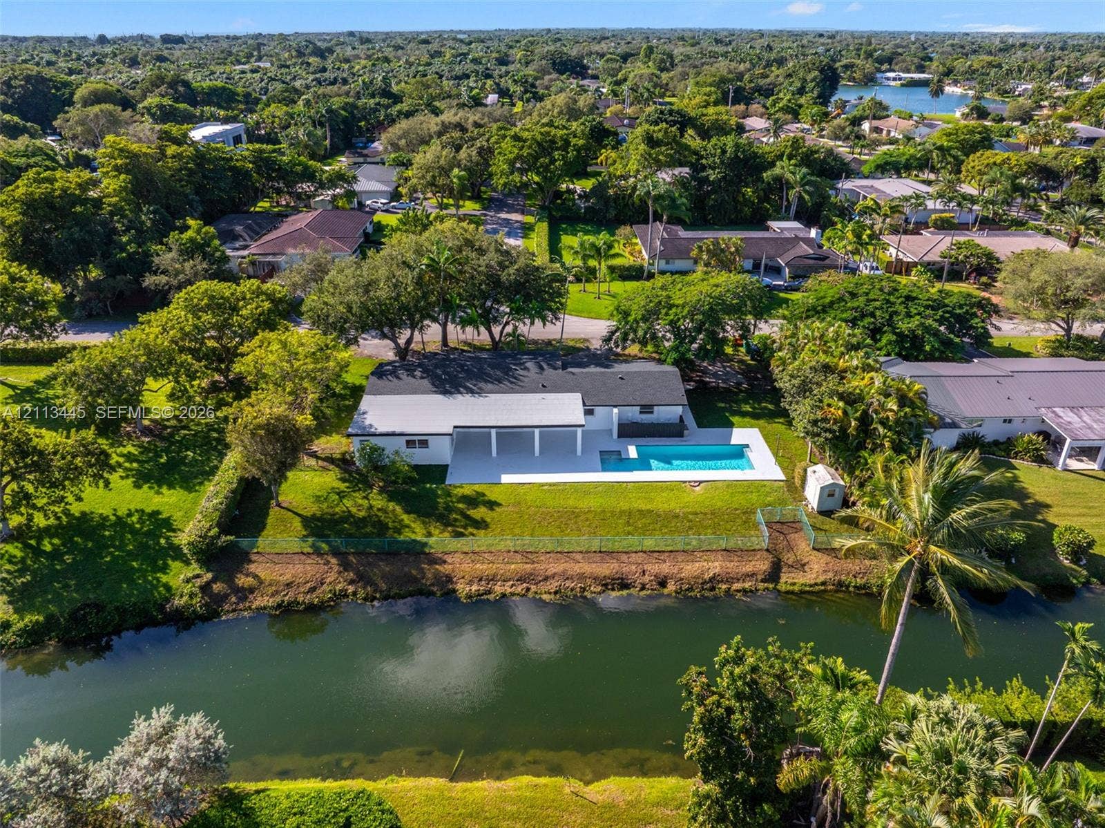 6951 SW 134th Street, Pinecrest, Pinecrest, FL 33156 Waterfront residence featuring a rectangular swimming pool, covered patio, sprawling lawn, and mature trees