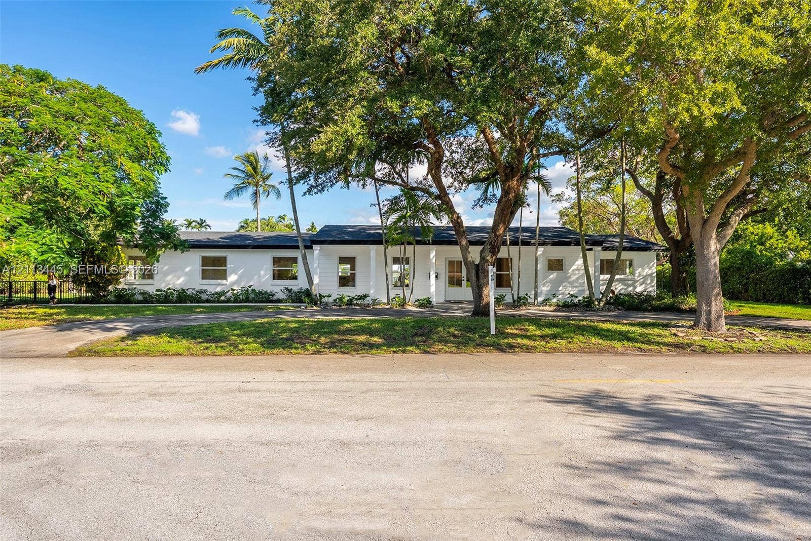 6951 SW 134th Street, Pinecrest, Pinecrest, FL 33156 Single-story residence featuring a white exterior, covered front porch, dark shingle roof, paved driveway, and mature landscaping