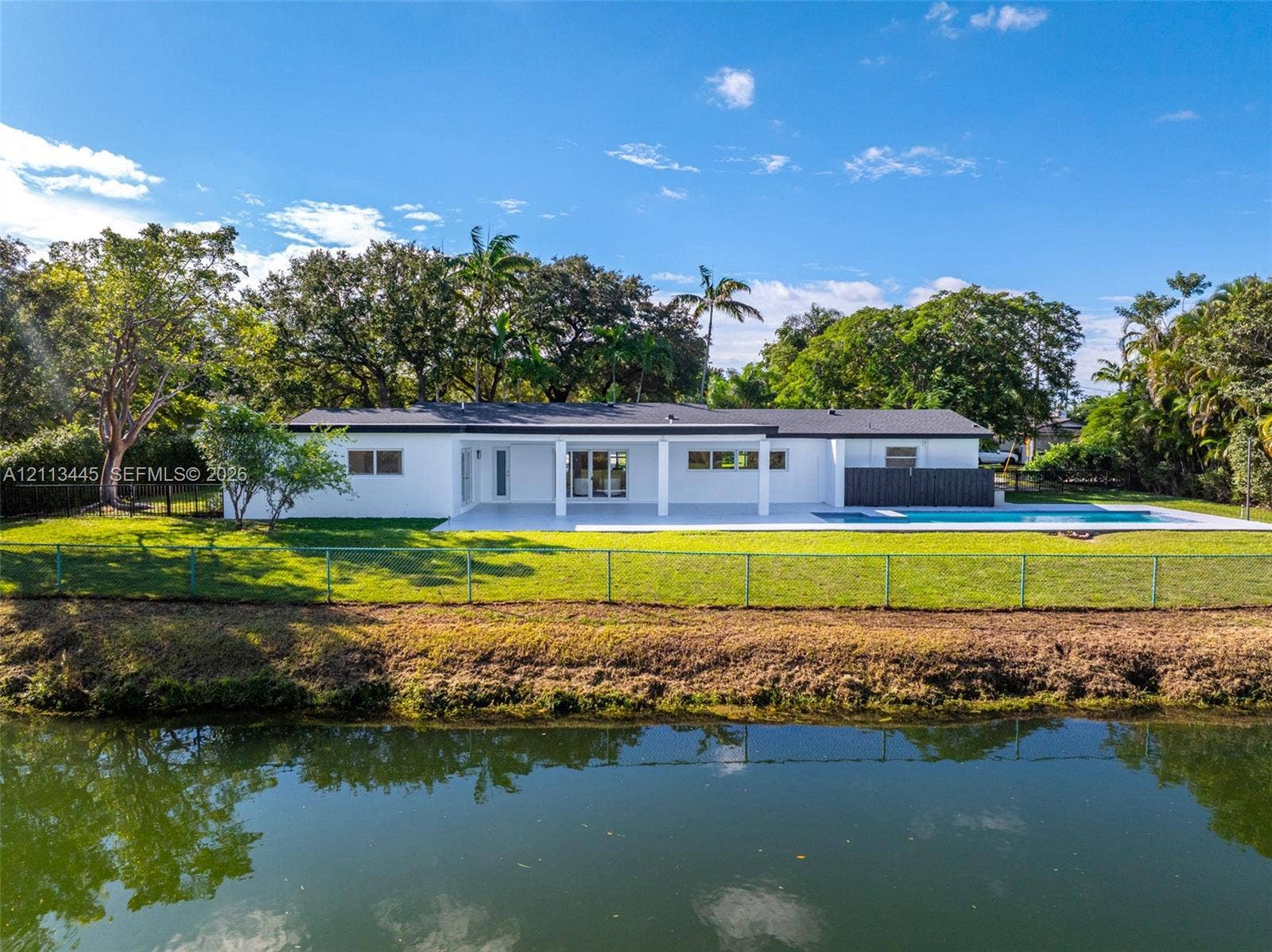 6951 SW 134th Street, Pinecrest, Pinecrest, FL 33156 Waterfront exterior featuring a covered patio, swimming pool, manicured lawn, and mature trees