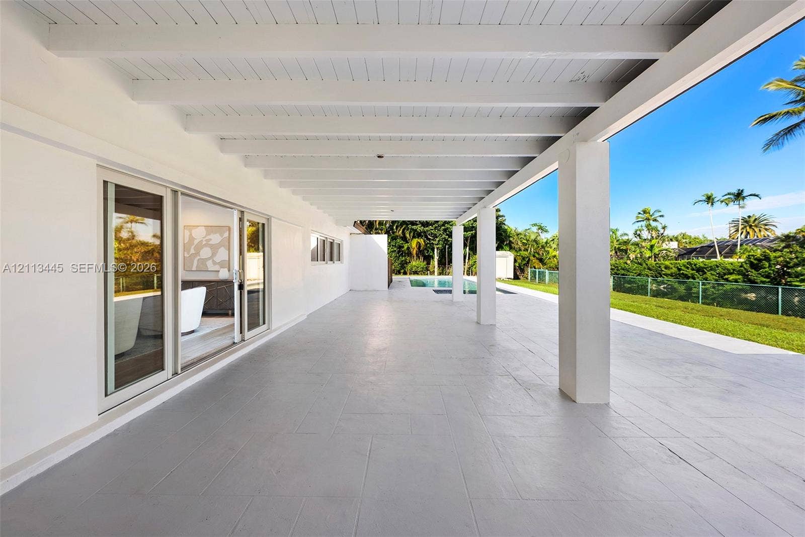 6951 SW 134th Street, Pinecrest, Pinecrest, FL 33156 Covered rear patio featuring a white-painted exposed beam ceiling, large-format tile flooring, and multiple sliding glass door entries