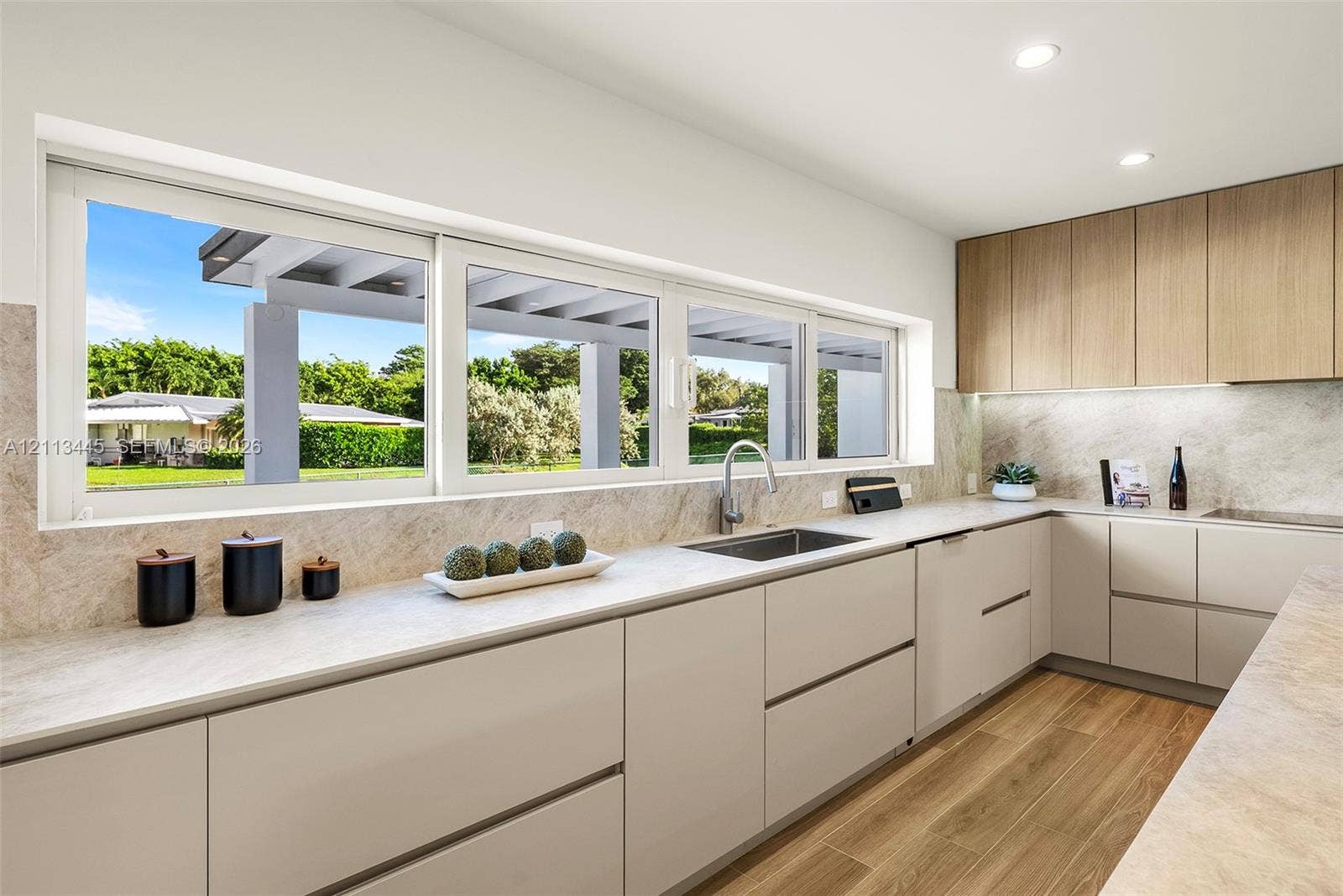 6951 SW 134th Street, Pinecrest, Pinecrest, FL 33156 Modern kitchen featuring light-toned cabinetry, wood-finish flooring, stone-textured countertops and backsplash, and an integrated undermount sink with a metallic faucet