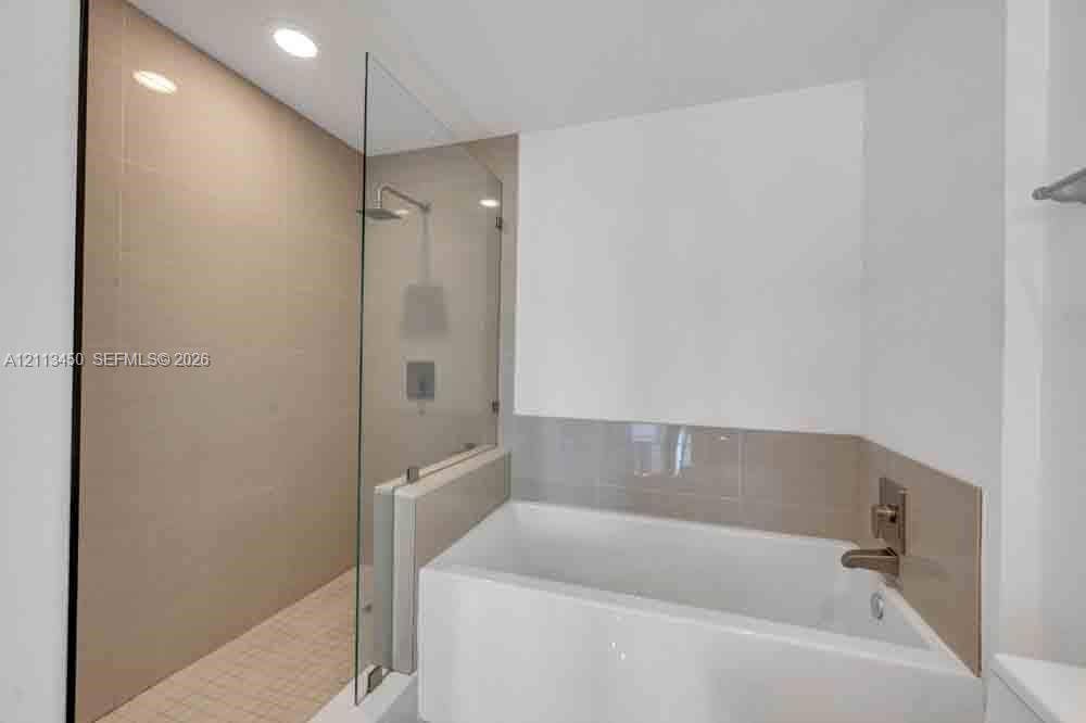 Walk-in shower with large-format wall tile, frameless glass enclosure, and ceiling-mounted rain showerhead, paired with a deep soaking tub featuring metallic fixtures and tile surround