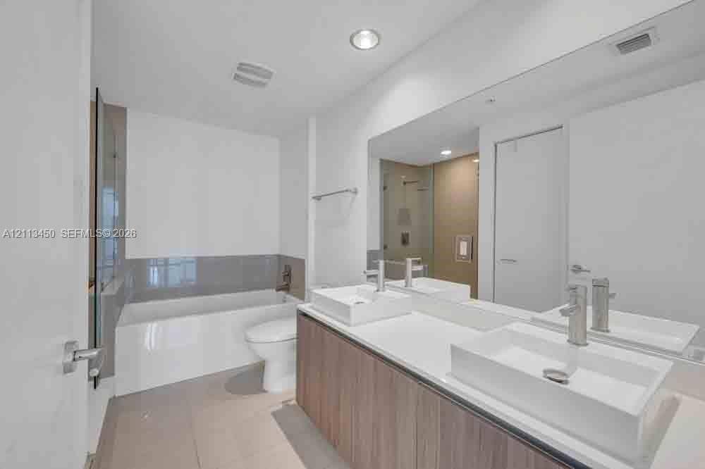Double vanity featuring wood-finish cabinetry, white solid-surface countertops, and rectangular vessel sinks with polished chrome faucets, complemented by a large wall-spanning mirror and a deep soaking tub with neutral tile surround