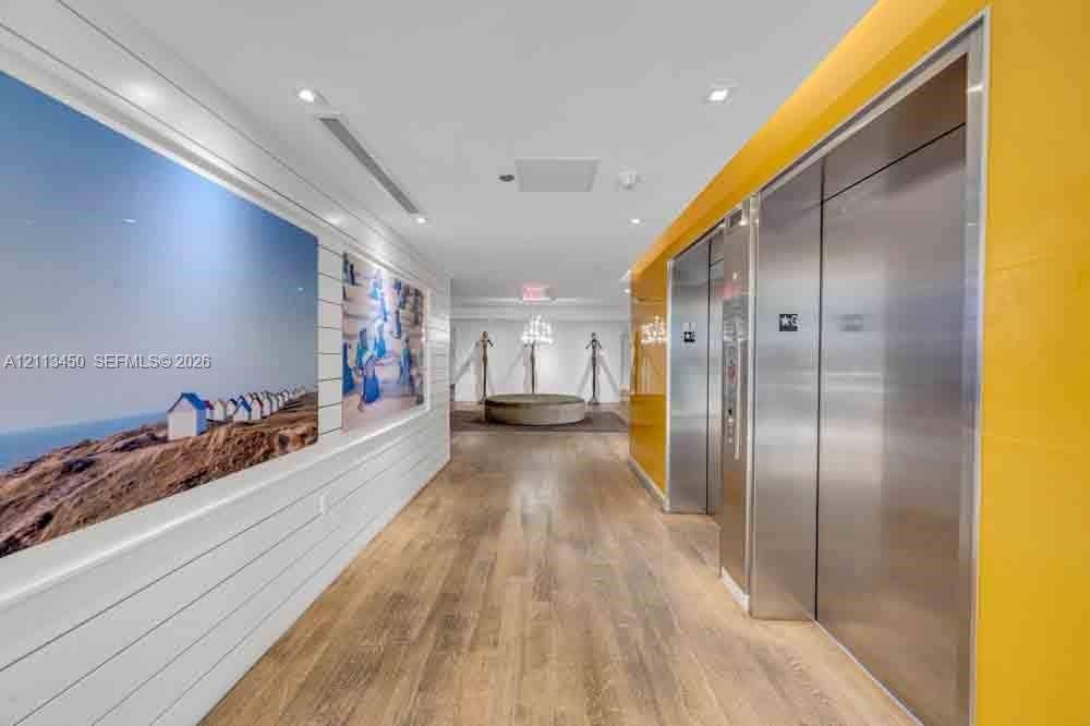 Common area corridor featuring wood-finish flooring, horizontal shiplap wall paneling, dual stainless steel elevator doors, and recessed ceiling lighting