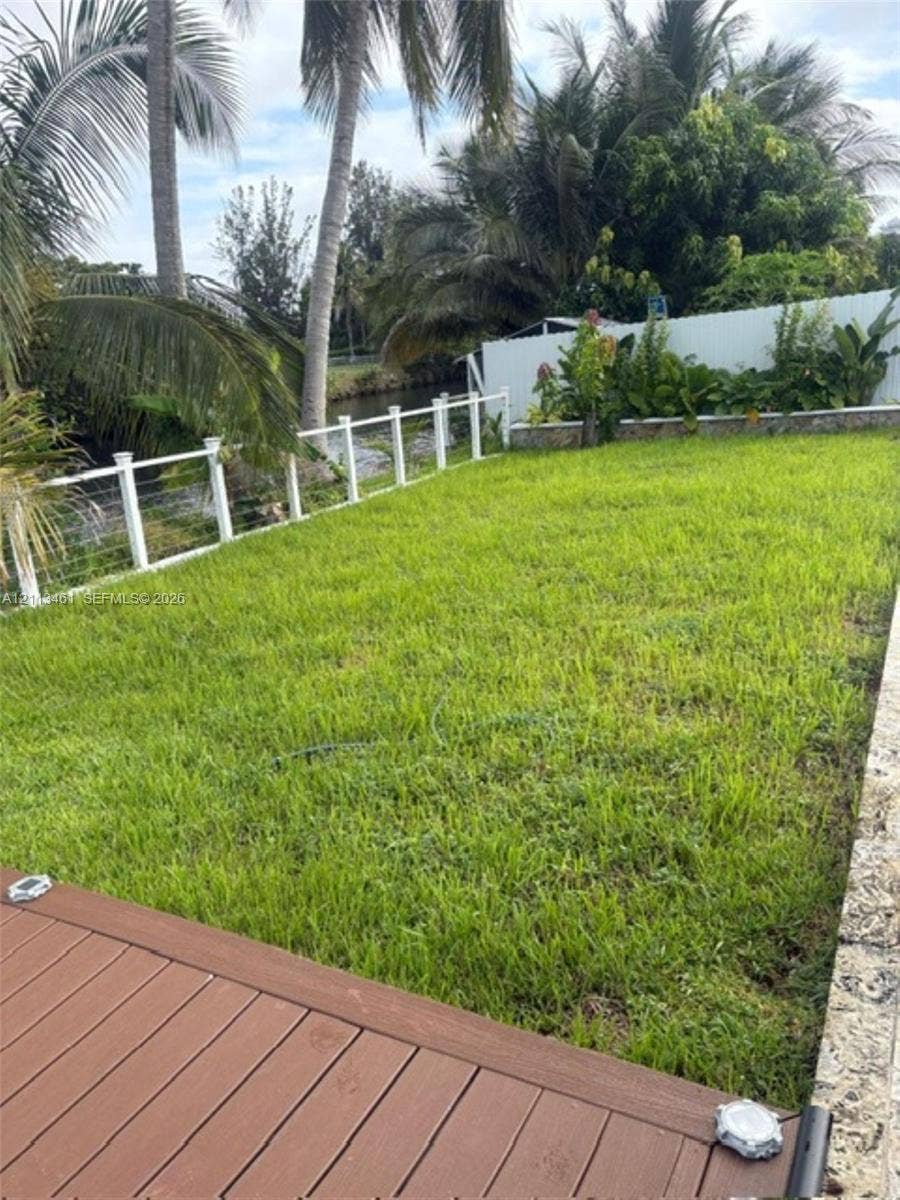 9961 SW 20th Street, Miami, Miami, FL 33165 Backyard featuring a wood-finish deck, natural grass lawn, white post-and-cable perimeter fencing, stone retaining wall, and mature palm trees